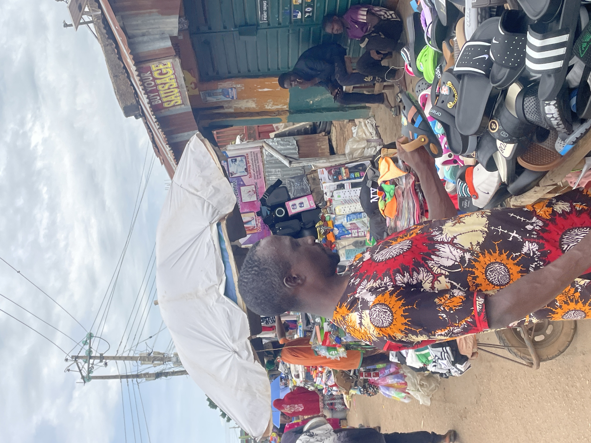 This is a street scene in Jos, Nigeria. The image captures an outdoor market where a man in a colorful shirt is examining a pair of sandals from a large display of footwear. He is standing at a stall with an abundance of sandals, flip-flops, and other foot-related accessories. The stall is covered by a white umbrella to provide shade.
In the background, another man is seated on a stool, looking down. Further back, a stall with a corrugated metal roof has a sign that reads "BUY YOUR SAUSAGE." Electrical poles and wires are visible against a cloudy sky. The ground is dirt and sand, typical of an unpaved market area. The overall impression is a bustling, informal marketplace.