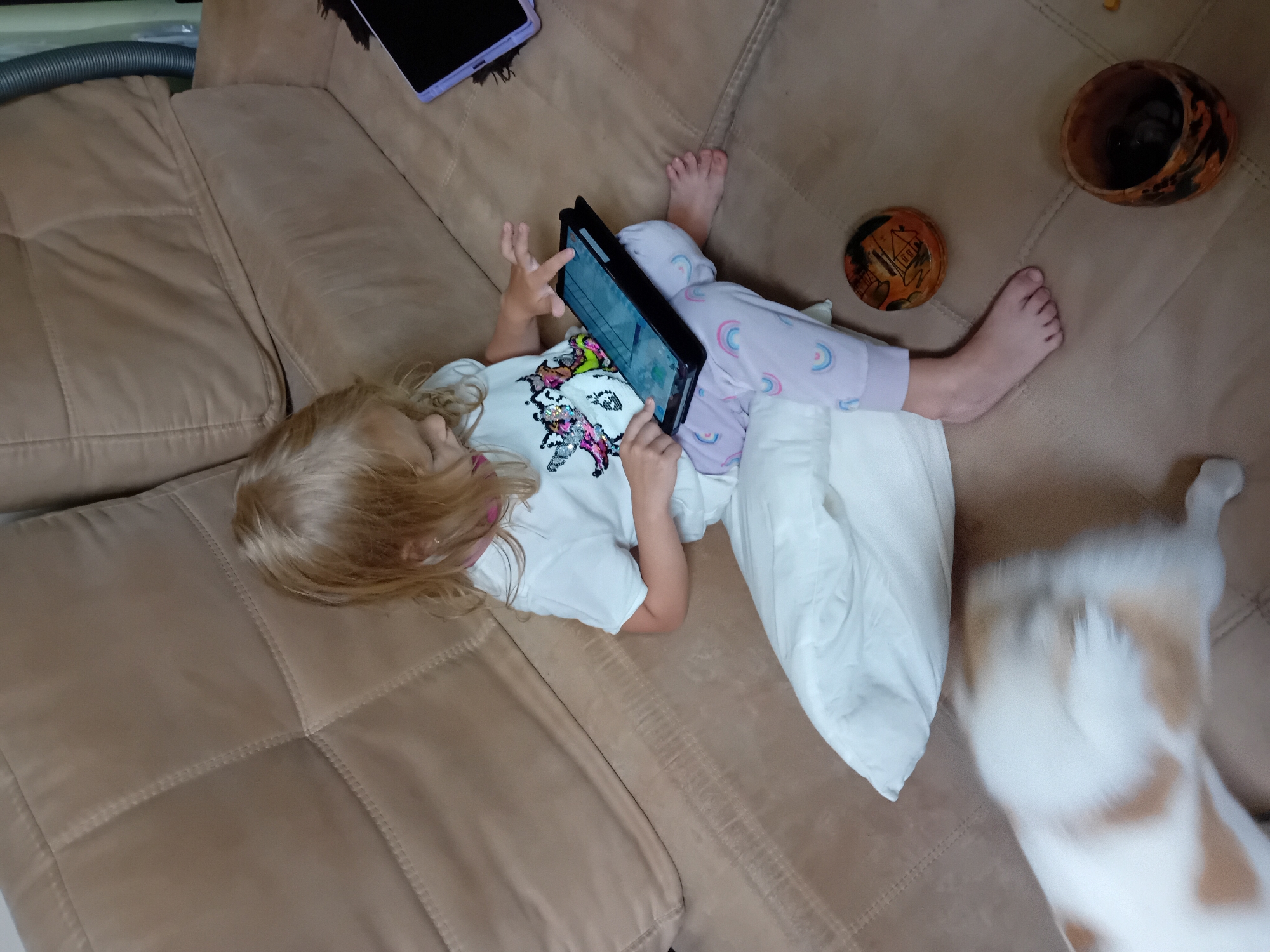 A young girl with blonde hair, wearing a white t-shirt with a unicorn graphic and light purple pajama bottoms with rainbow patterns, is sitting on a beige upholstered couch. She is holding a black tablet in her lap and appears to be interacting with it, with her right index finger extended towards the screen. A white pillow is positioned behind her, and to her right, on the couch, is another tablet with a purple case. In the foreground, a white and orange-spotted cat is partially visible, blurred due to motion. Two decorative wooden bowls, one open and containing dark objects, are on the couch near the girl's feet. The scene suggests an indoor domestic setting, likely a living room. There is no information in the image to suggest a location in Chuhuiv, Ukraine.