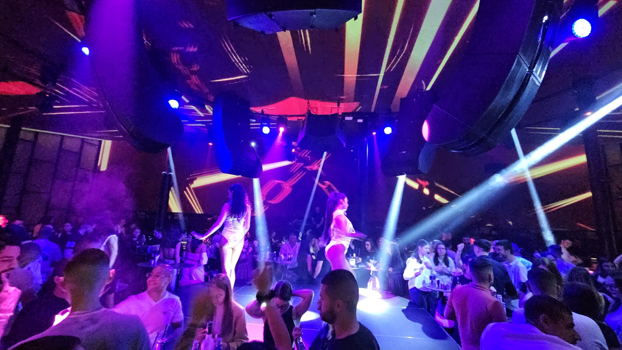 The image shows a crowded nightclub in Varna, Bulgaria. Two women in white outfits are performing on a raised platform under blue and red stage lights. Laser beams cut through the air. The audience is seated at tables, drinking, and watching the show. Large speakers hang from the ceiling above the stage. The ceiling is decorated with illuminated stripes.