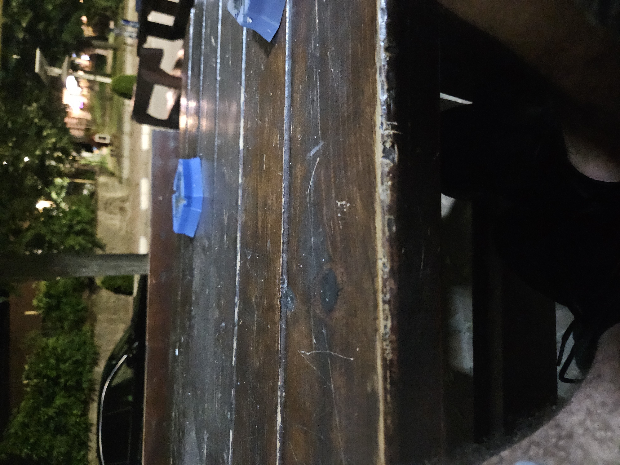The image depicts an outdoor setting, likely a cafe or restaurant patio, at night. A dark wooden table occupies the foreground, showing visible wear and scratches. On the table, a blue ashtray is visible on the left, and a portion of another blue object, possibly another ashtray or part of a seating area, is seen on the right. In the background, trees with foliage are illuminated by ambient light, suggesting streetlights or building lights. A dark-colored sedan is parked on the left side of the frame, partially obscured by the table. The overall scene is dimly lit, with a focus on the textured surface of the table. The mention of Biaga, Bulgaria, is external context and not visually verifiable from the image itself.