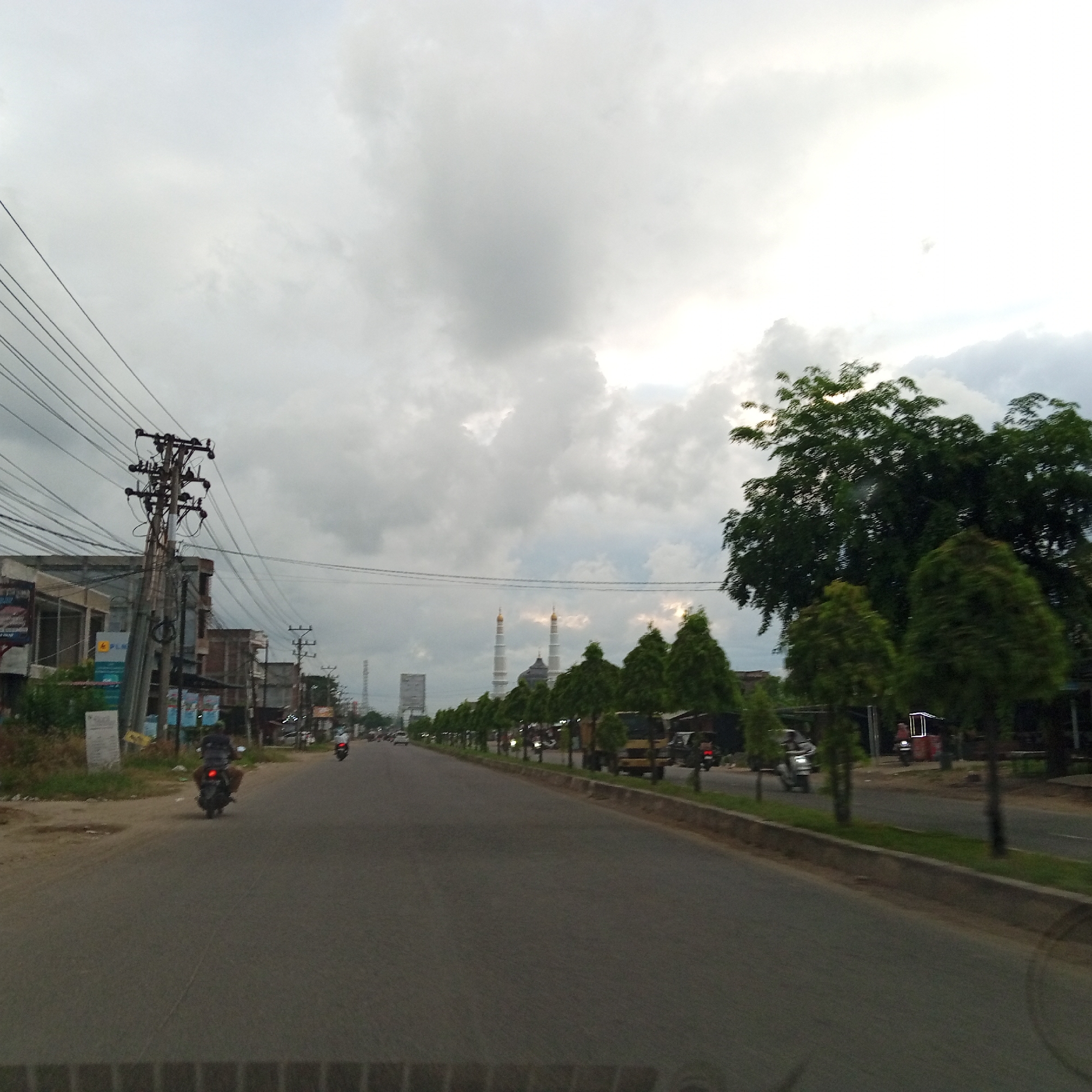 This image depicts an overcast street scene in Pantegajah, Indonesia, captured from a moving vehicle. The sky occupies the upper half, dominated by thick, gray clouds with brighter areas suggesting diffuse sunlight on the right.
In the foreground, the asphalt road surface is visible, dark gray and appearing slightly damp or textured. There are no clear lane markings.
On the left side of the road, a dense network of black electrical or communication wires spans the upper left quadrant, attached to a prominent utility pole with multiple cross-arms and insulators. Below the wires, a row of two-story buildings lines the street, some appearing to be shops or residences, constructed from concrete or brick. A sign displaying "PLN" (Perusahaan Listrik Negara, Indonesian state electricity company) is visible on one of these buildings. A small white informational sign stands on the unpaved verge in front of the buildings. A motorcyclist in dark attire is visible on the road, heading away from the camera, roughly in the center-left. Other motorcycles are visible further down the road on the left side.
On the right side of the road, a raised concrete curb separates the asphalt from a strip of green grass and a continuous row of neatly trimmed, conical evergreen trees. Behind these smaller trees, larger, irregularly shaped trees with dense green foliage are visible. Further down the road, behind the trees, various vehicles including a light-colored truck and a three-wheeled vehicle with an illuminated frame can be discerned.
In the mid-distance, the most prominent features are two tall, slender white mosque minarets topped with golden domes, positioned centrally in the frame. A darker dome structure of the mosque is situated between and slightly in front of the minarets. To the left of the mosque, a tall, slender telecommunication tower or mast is visible on the horizon, alongside a large, rectangular billboard structure.
The overall environment suggests a semi-urban setting, characterized by mixed commercial and residential structures, overhead utility lines, and common road traffic, predominantly motorcycles. The cloudy sky casts a soft, uniform light over the entire scene.