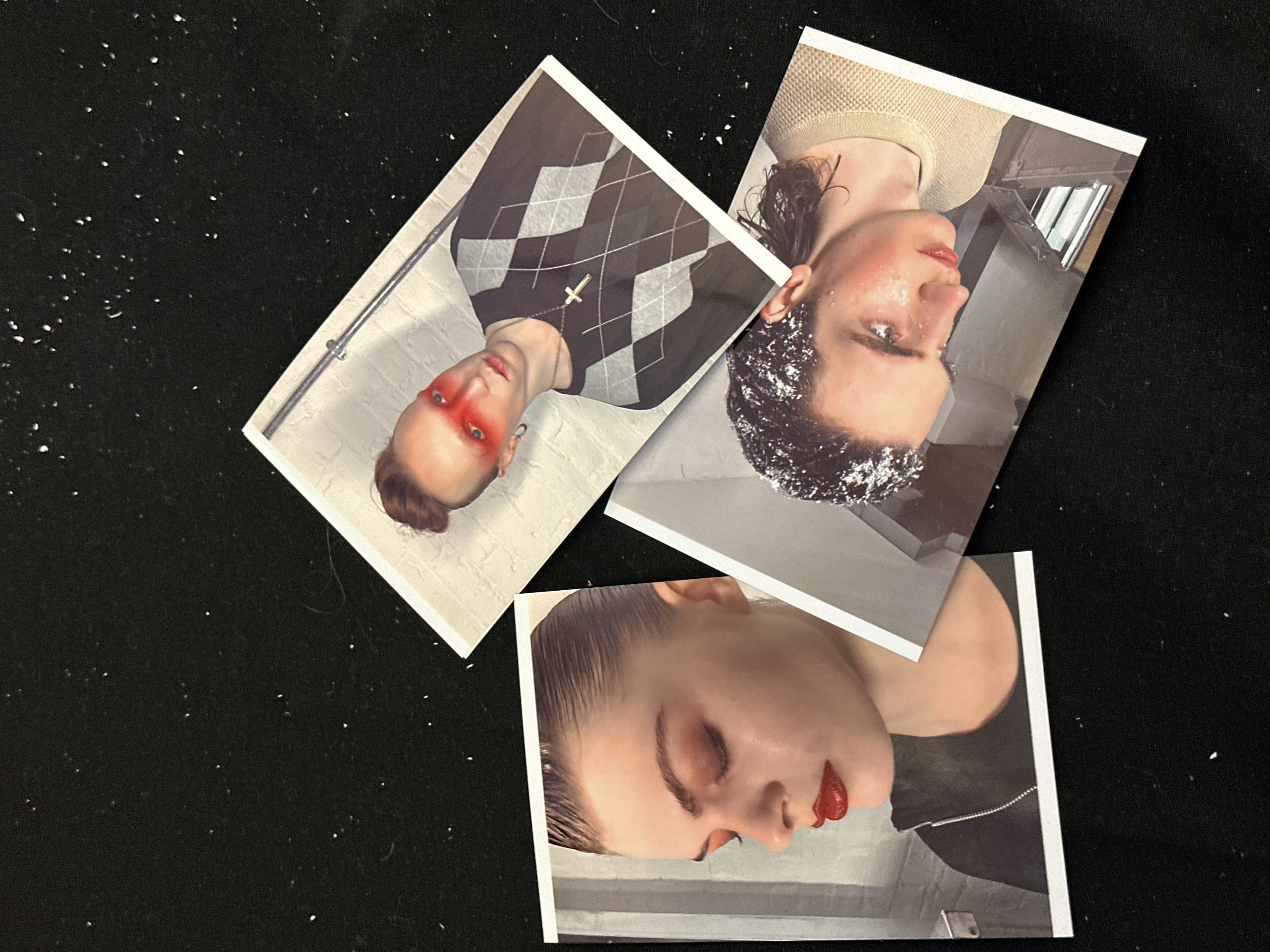 Three printed photos sit on a black surface speckled with tiny white specks. One features a person with red makeup around their eyes, wearing an argyle sweater and a cross necklace. Another shows a person with glitter on their hair and face, wearing a tan top. The third photo depicts a person with closed eyes and red lips. The prints overlap, creating an informal collage effect.