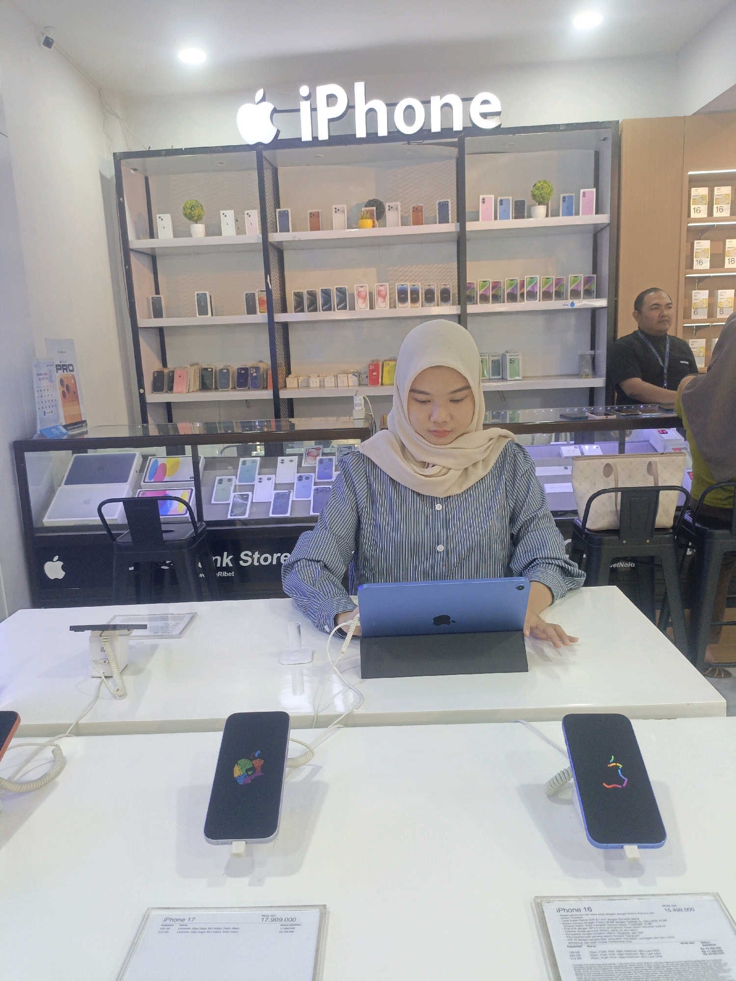 In a well-lit, modern retail store, likely an electronics shop, a young woman wearing a light beige hijab and a blue and white striped long-sleeved shirt is seated at a white counter, looking at a blue tablet in front of her. She appears to be interacting with or demonstrating the tablet, as her right hand is resting near it. The store's interior features display shelves stocked with various electronic devices, including numerous iPhone boxes and a few decorative plants. Above the shelves, a prominent "iPhone" logo is illuminated. In the background, to the right, a man in a black polo shirt with a lanyard is standing, possibly a store employee, and partially visible to his right is another person. The counter in the foreground displays two iPhones, one silver and one purple, both on display stands and showing a colorful Apple logo on their screens. Below the iPhones are informational displays with text in Indonesian, indicating product names like "iPhone 17" and "iPhone 16" along with prices. The overall environment suggests a retail setting for Apple products. This scene is not specifically identifiable as being located in Probolinggo, Indonesia, based solely on the visual information provided.