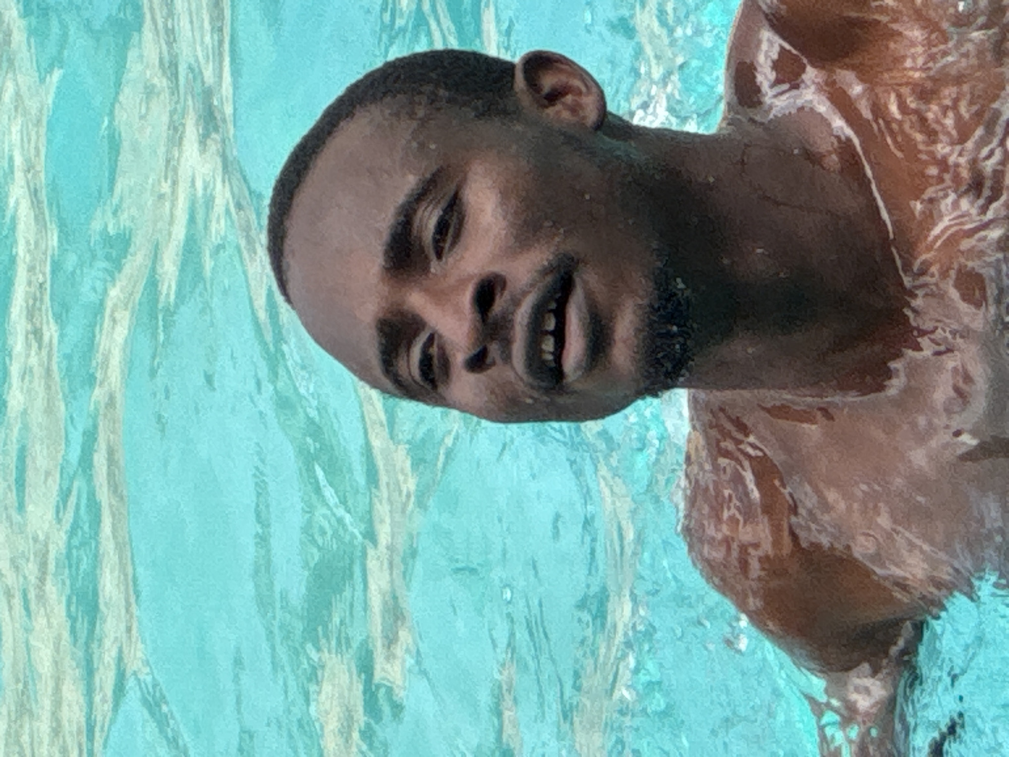 A close-up, waist-up shot shows a dark-skinned man partially submerged in bright blue water. The water appears to be in a pool, with sunlight creating ripples and highlights on the surface and on the man's skin. He is looking upwards and to his left, with his mouth slightly open, revealing his teeth. He has a short beard and his head is shaved. The man's shoulders and upper chest are visible, glistening with water. The background is entirely composed of the textured, turquoise water. The image captures a moment of leisure, possibly in a swimming pool in Nkpor, Nigeria, given the context provided, though the image itself does not contain any explicit indicators of this location.