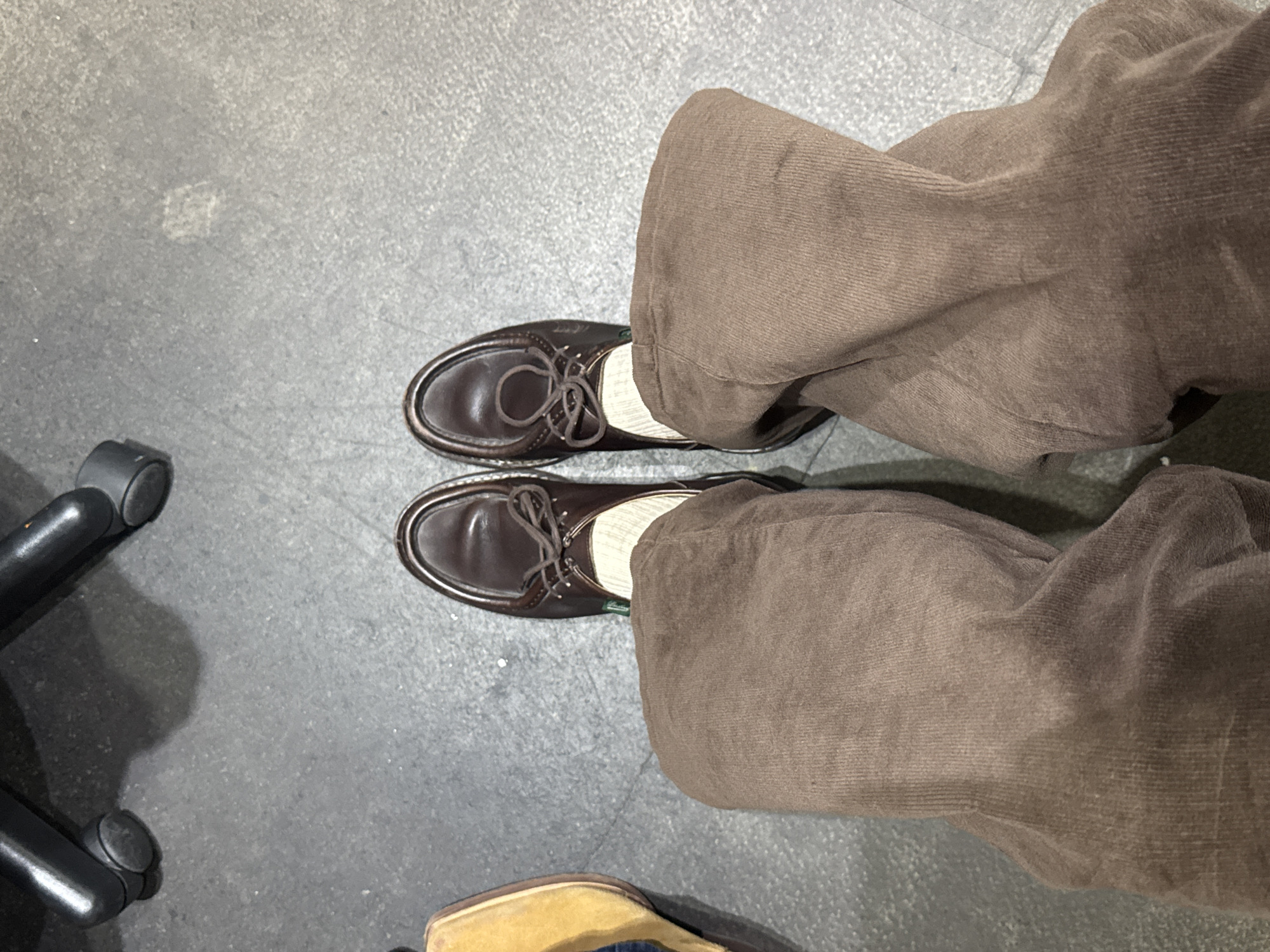 The image is a top-down view of a person's legs and feet. The person is wearing brown corduroy pants and brown leather shoes with white socks. The person is sitting in a chair on a gray concrete floor. The location is not identifiable within the city of London, United Kingdom, based on the information in the image.