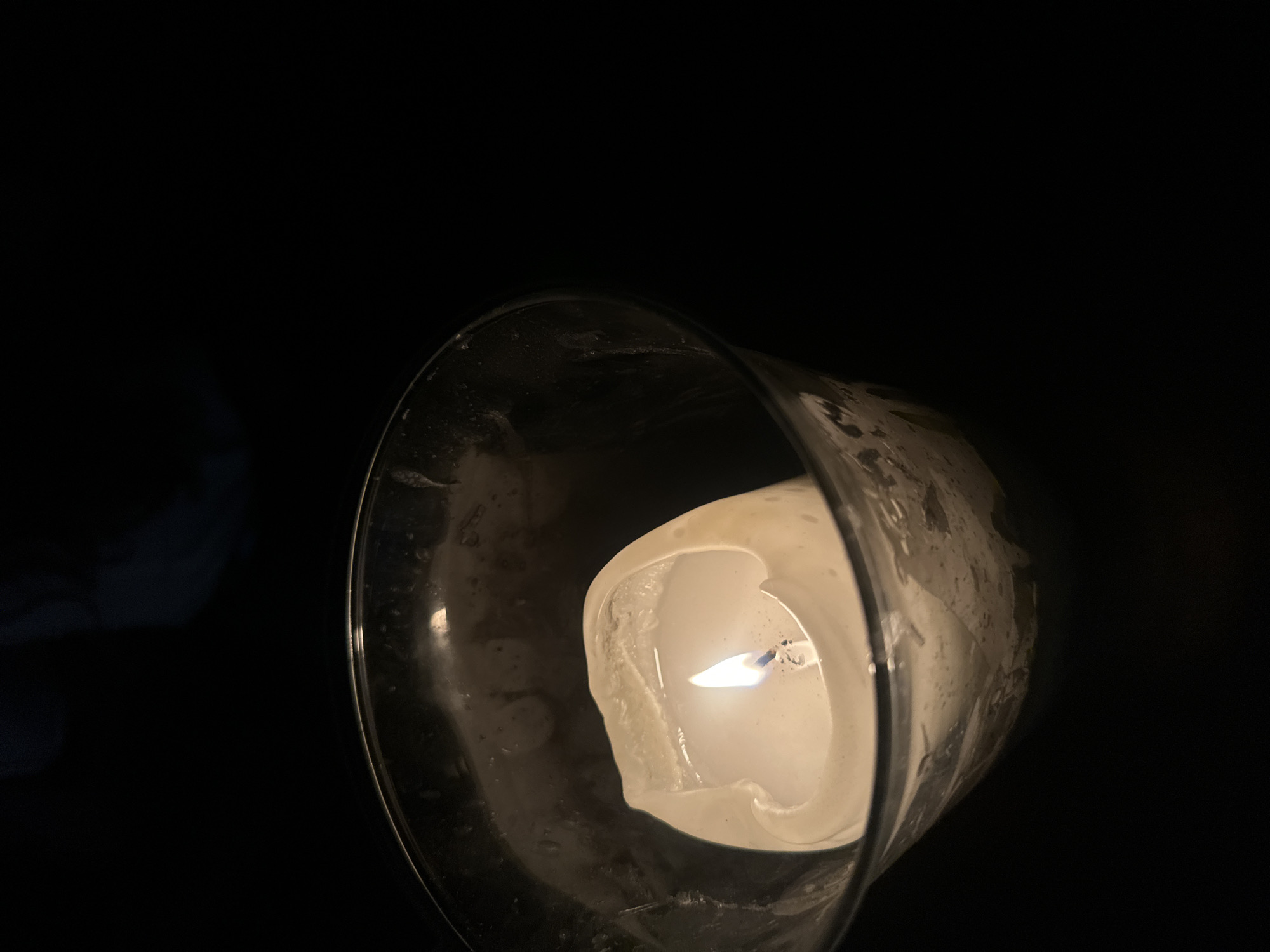 The image shows a lit candle inside a glass holder, positioned against a dark background. The candle is partially melted, and the flame is visible. The glass holder appears to be somewhat frosted or has residue on its surface. The scene is dimly lit, with the candle providing the primary source of illumination. The location is not discernible from the image.