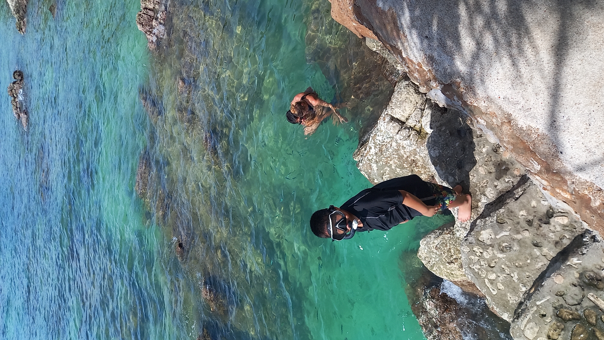 The image captures a scene on the coast of Koh Phangan, Thailand. In the foreground, a young boy wearing a black rash guard and snorkel mask stands on a rocky surface, facing the camera. He is barefoot and has colorful patterned shorts. In the background, two individuals, a man and a woman, are in the shallow, turquoise-colored ocean. The man has his arm around the woman. The water is clear, revealing rocks and small fish beneath the surface. The environment is coastal with a clear sky and sunlight illuminating the scene.