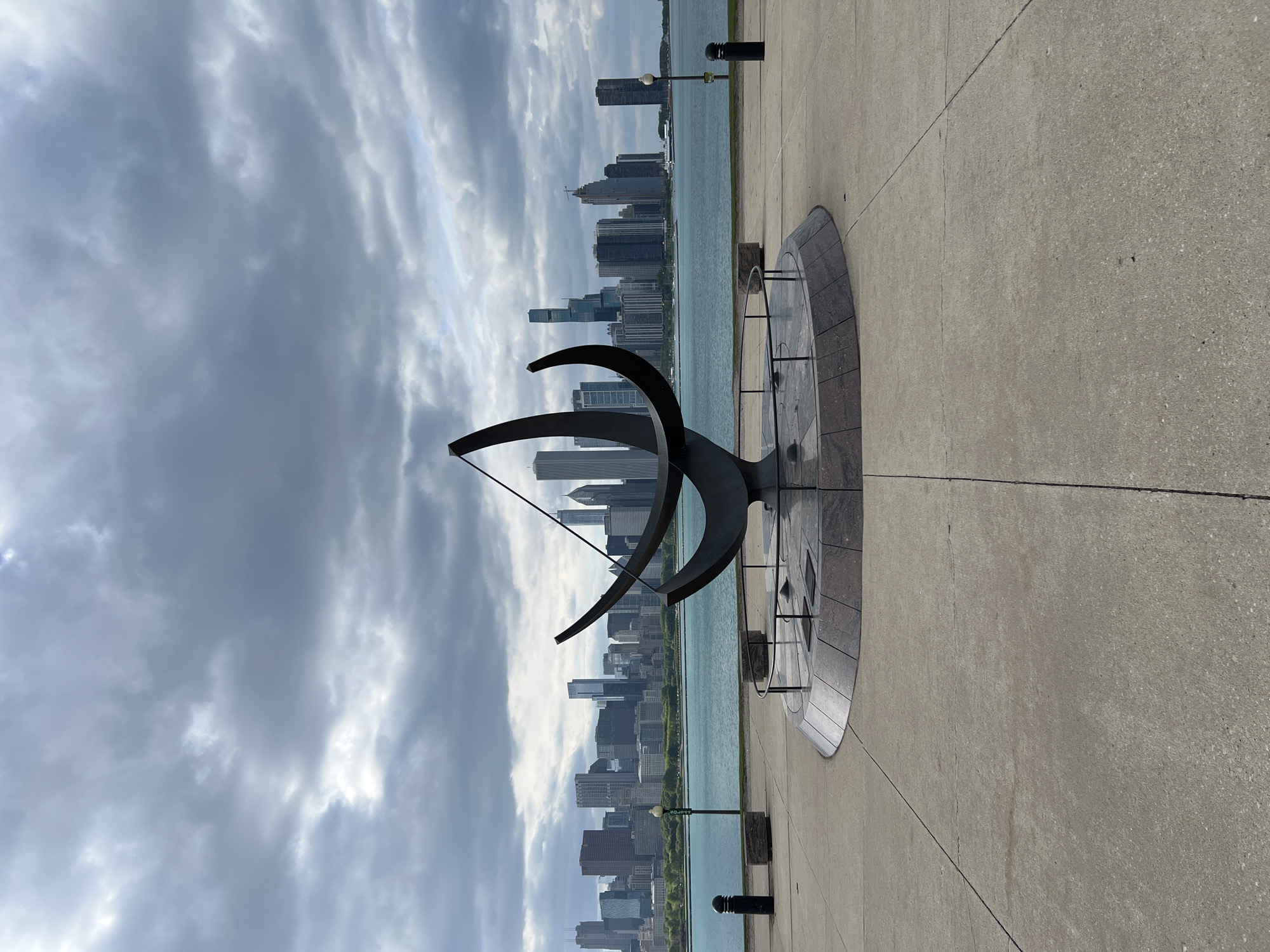 The image shows a large sundial sculpture on a concrete platform, with the Chicago skyline and Lake Michigan in the background. The sky is overcast with dark clouds. There are no people visible in the image.