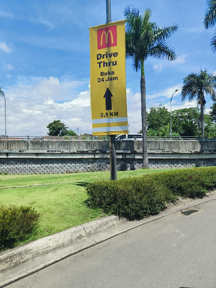 The image shows a yellow sign with the McDonald's logo on it. It says "Drive Thru Buka 24 Jam 2,5 KM" indicating a drive-thru McDonald's restaurant is 2.5 kilometers away and open 24 hours a day. The sign is attached to a metal pole in a grassy area. The pole is situated between a concrete wall and a paved road. The area is well-maintained and features lush green bushes and grass. The location is in the city of Padalarang, Indonesia, at coordinates -6.852 latitude and 107.496 longitude. There are no people or vehicles in the image. The sky is blue with white clouds.
