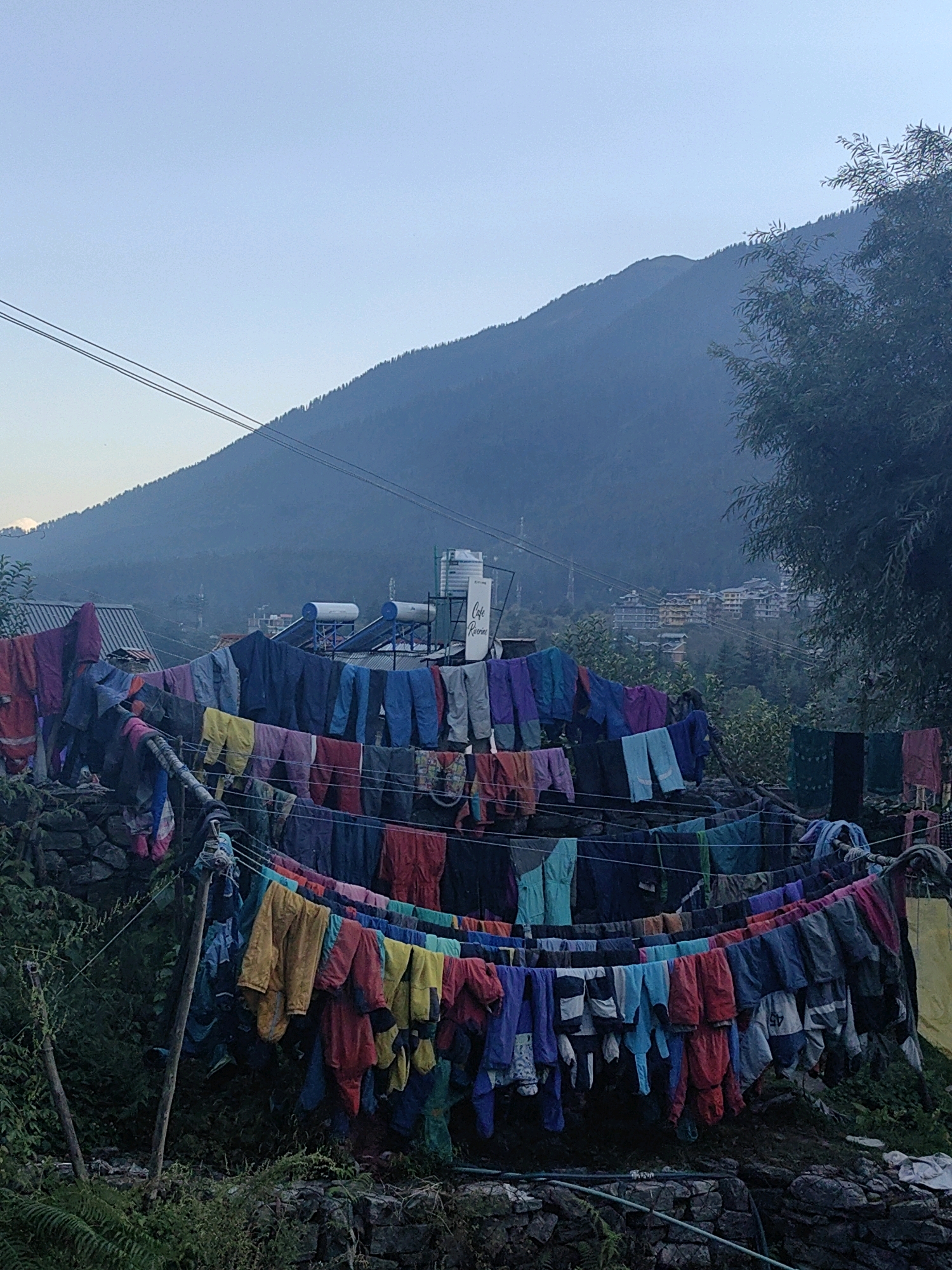 This photograph depicts a scene in Vashisht, India, where laundry is hung to dry on multiple clotheslines strung across a hillside. The clothes are a vibrant mix of colors, including reds, blues, yellows, purples, and greens, and appear to be various articles of clothing such as pants, shirts, and jackets. The laundry is supported by wooden poles and wires, and some of it hangs over a stone wall.
In the background, a misty mountain range rises, with trees dotting its slopes. A few buildings, likely part of the town, are visible nestled on the mountainside, some appearing to be residential or commercial structures with solar water heaters on their roofs. The sky above is a pale, hazy blue, suggesting either early morning or late afternoon light. The overall impression is one of everyday life in a mountainous region, with the colorful laundry adding a striking visual element against the natural landscape. No people are visible in the frame.