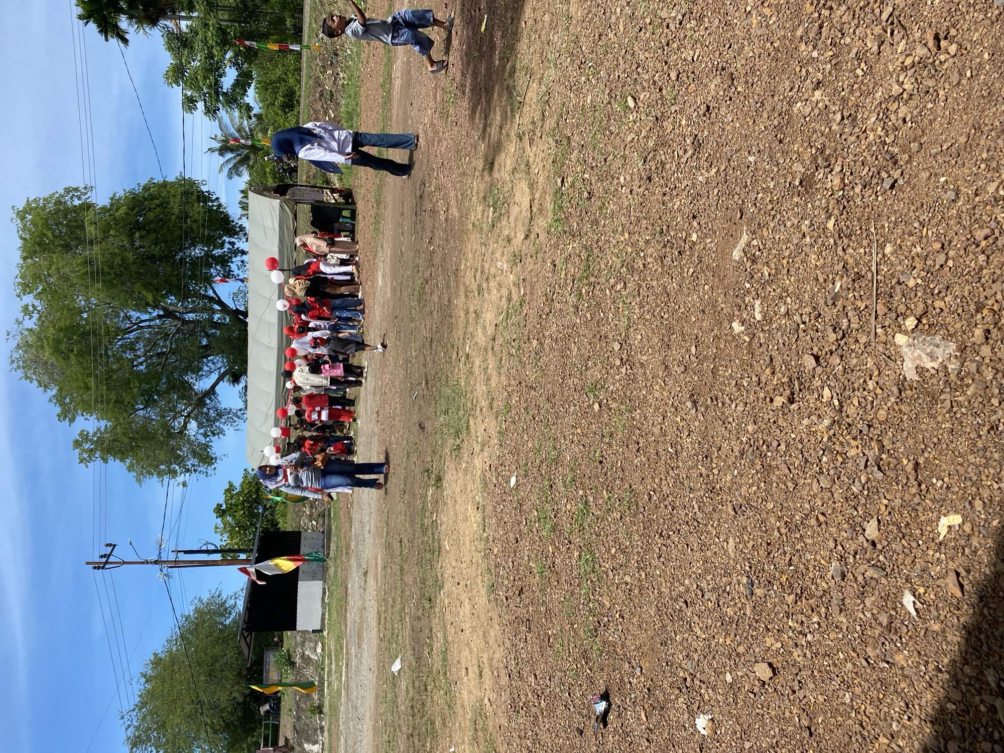 A group of people, including men, women, and children, are gathered outdoors in a dusty, gravelly area. Many are wearing red and white, suggesting a celebratory or patriotic event. Some are standing under a large tent, while others are milling about in the open. In the background, there are trees, power lines, and a small building with flags. One woman in the foreground, wearing a blue hijab and jeans, is walking towards the viewer. A young boy in a grey shirt and blue shorts is running in the mid-right of the frame, looking towards the left. The scene appears to be in a rural or semi-rural setting in Aneuk Paya, Indonesia.