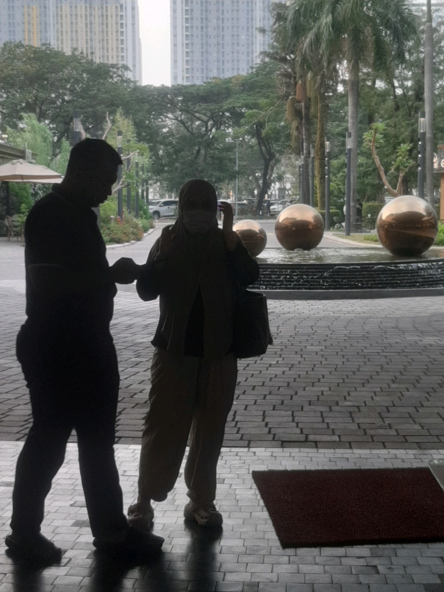 A man and a woman are standing outside a building, near a water feature with three large golden spheres. The man is on the left, silhouetted and wearing dark clothing. The woman is on the right, wearing a headscarf and a light-colored outfit, with a dark bag on her shoulder. They appear to be interacting or talking. In the background, there are tall buildings, trees, and palm trees, suggesting an urban environment. The ground is paved with gray and white bricks. The overall scene suggests a public space, possibly a hotel or a commercial area, located in the city of Bekasi, Indonesia.