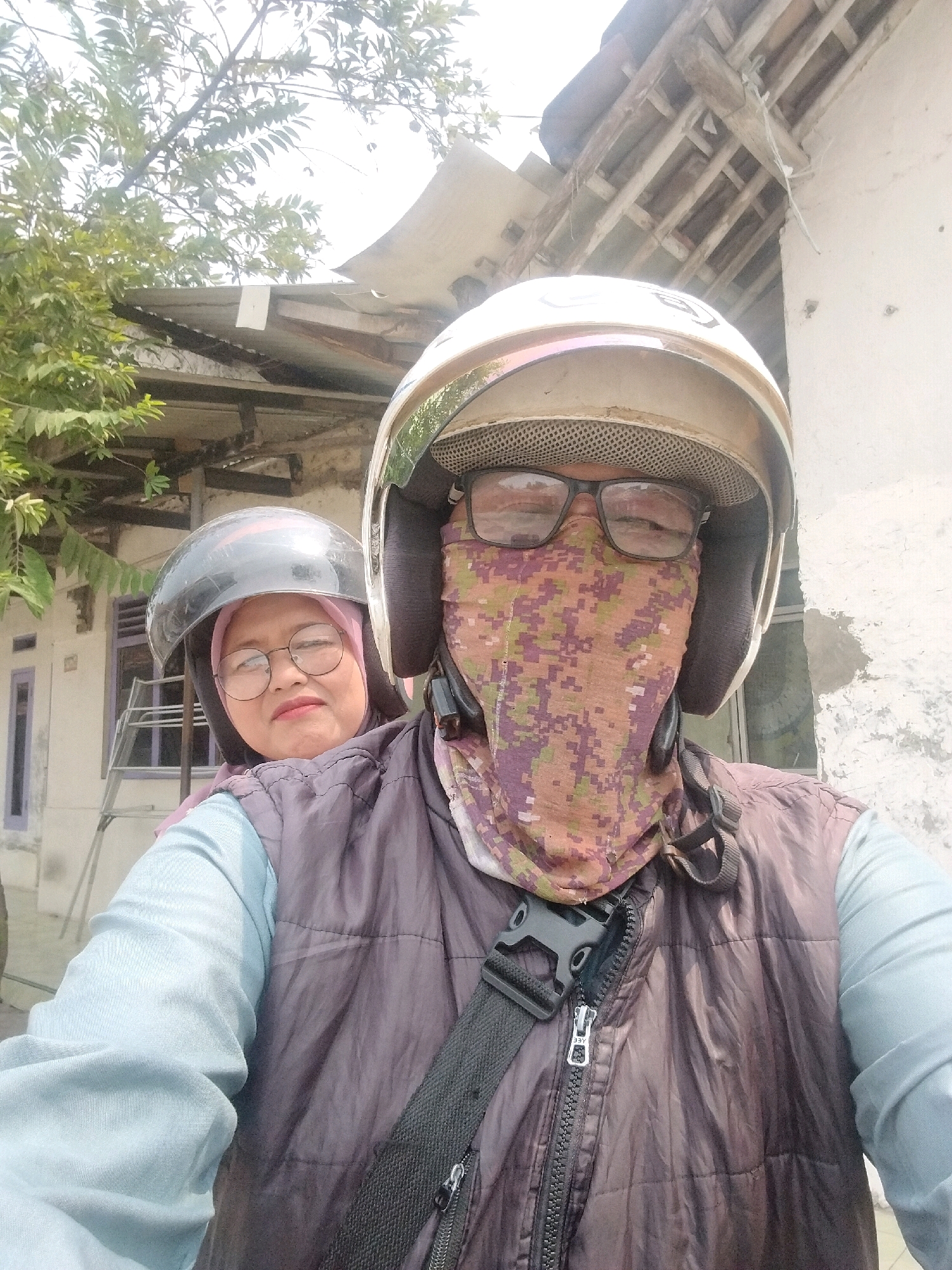 In the city of Serang, Indonesia, a selfie is taken from the perspective of someone riding a motorcycle. The rider, wearing a white helmet, glasses, and a camouflage mask, is in the foreground. Behind them, a woman wearing a black helmet and glasses is visible. The background shows a building with a rustic tiled roof and some greenery. The lighting suggests it is daytime.