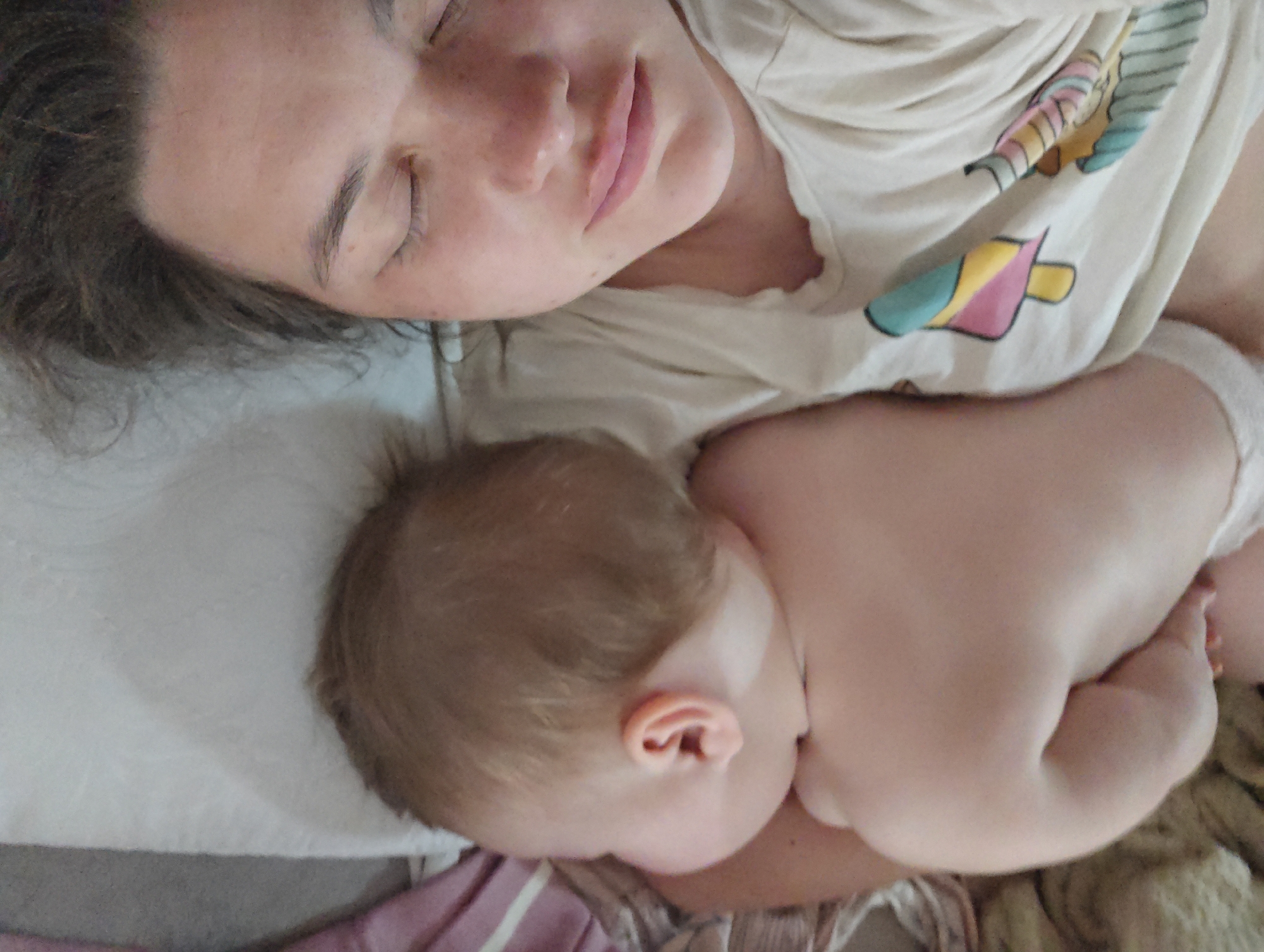 A woman and a baby are lying down. The woman is on her back, with her eyes closed, and a light-colored patterned t-shirt is visible. The baby is lying next to her, facing away from the camera. The baby has fair skin and brown hair and is wearing a white diaper. They are lying on bedding consisting of a white sheet, pink striped fabric, and beige fabric. The location is unknown, but is stated to be Chaikovsky, Russia.