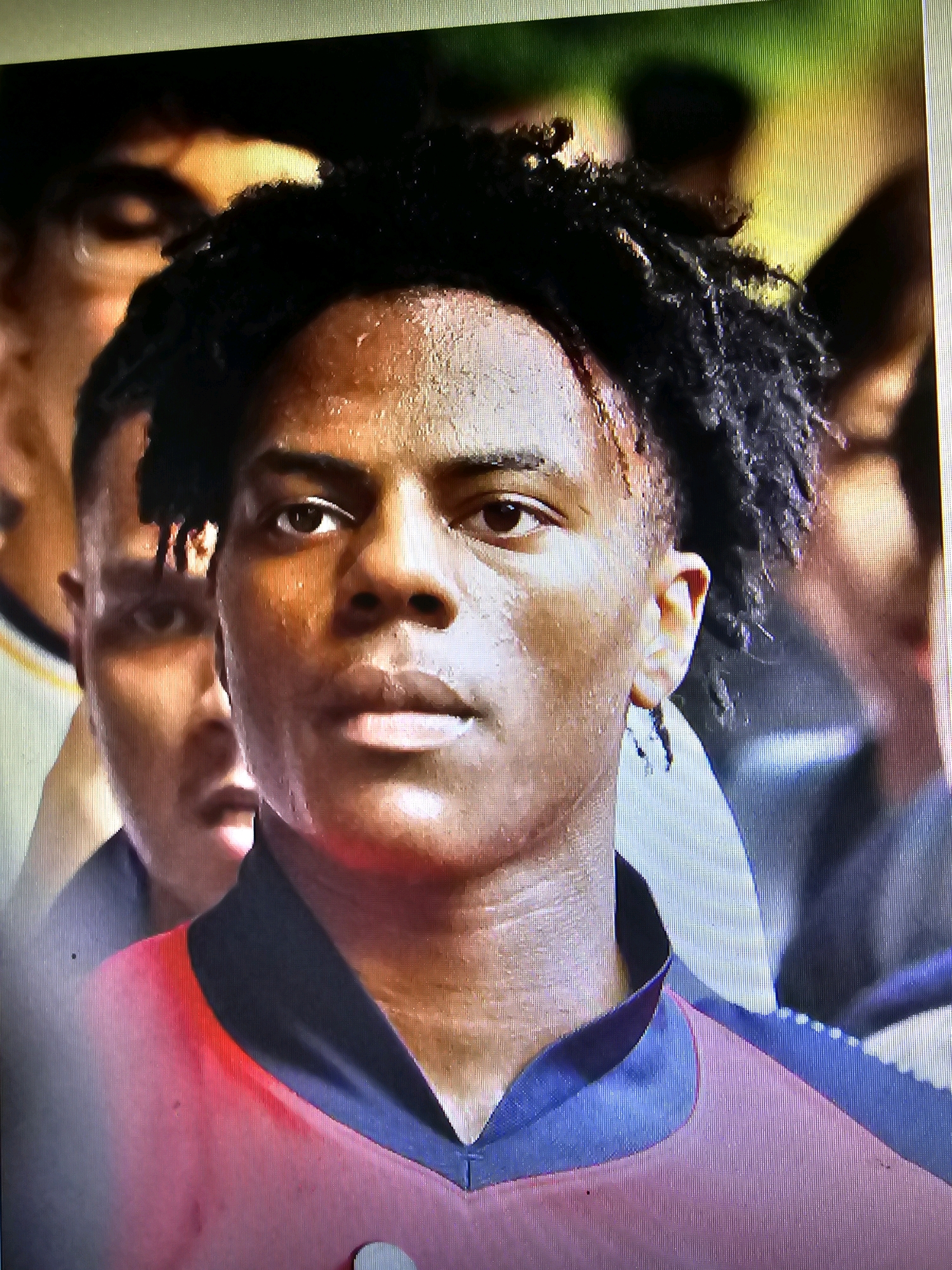 The image is a close-up, slightly blurry photograph of a young Black male with dark, textured hair, facing slightly to his left. He has sweat glistening on his forehead and cheeks, indicating exertion or heat. His eyes are open, looking forward, and his mouth is slightly parted. He is wearing a collared garment that is red with blue trim. In the background, two other individuals are partially visible, also appearing to be young males, with one directly behind and to the left of the main subject. The lighting is somewhat dim, with a warm glow on the faces of the subjects. The overall impression is that of a candid or documentary-style photograph, possibly taken during an athletic event or a similar activity, given the apparent sweat and the presence of other people. There is no indication of the city of Batna, Algeria, within the image itself.