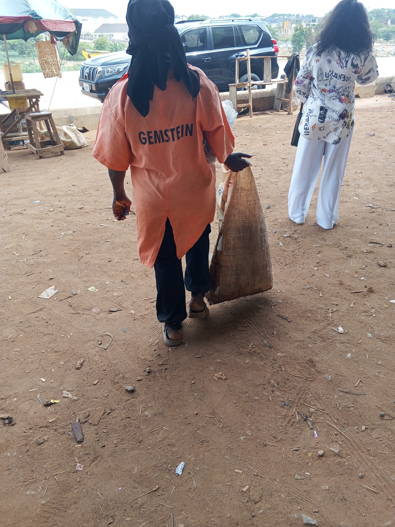 The image shows an outdoor scene in Galadimawa Village, Nigeria. A person wearing an orange shirt with the word "GEMSTEIN" on the back, black pants, and sandals is carrying a large sack. Another person in a white patterned shirt and white pants stands nearby. The ground is dirt and debris-strewn. In the background, there is a black SUV, a simple wooden structure, and a building. A partial view of a vendor's stall is also visible.