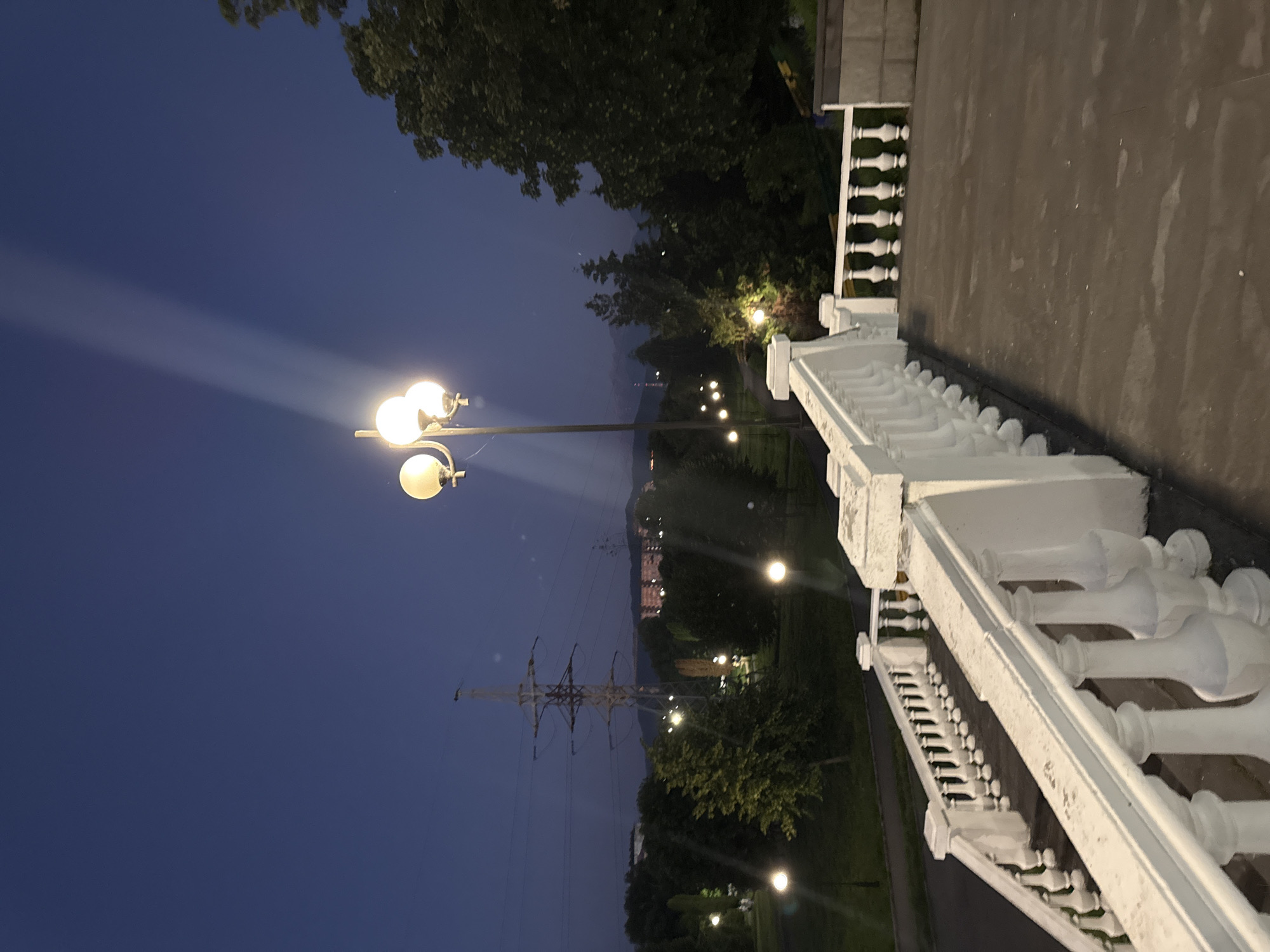 The image depicts a nighttime scene in Arkabash 1-Y, Russia. The foreground features a white decorative railing, characteristic of a park or public walkway. Beyond the railing, a path curves through a park area, illuminated by several lampposts that cast pools of light. In the mid-ground, a large transmission tower stands tall, its structure outlined against the dark blue twilight sky. Trees dot the landscape, their leaves appearing dark and dense in the low light. In the distance, faint lights suggest buildings or a settlement nestled among rolling hills or mountains. The sky is a deep twilight blue, indicating that it is either early evening or late evening. The overall mood is serene and quiet, with the artificial lights providing a warm contrast to the fading daylight. There is no visible text in the image.