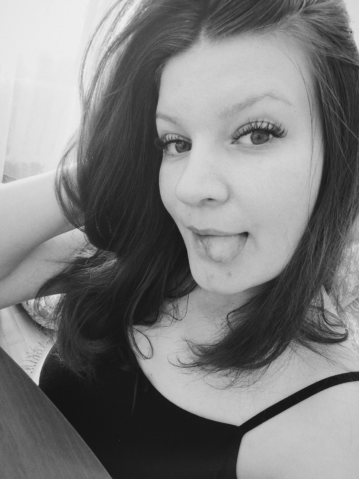 A black and white portrait shows a woman with long dark hair, wearing a black tank top. She has her hand behind her head and is sticking her tongue out. The background is blurred, suggesting an indoor location near a window with diffused light. The image does not provide enough detail to identify a specific location in Tobolsk, Russia.
