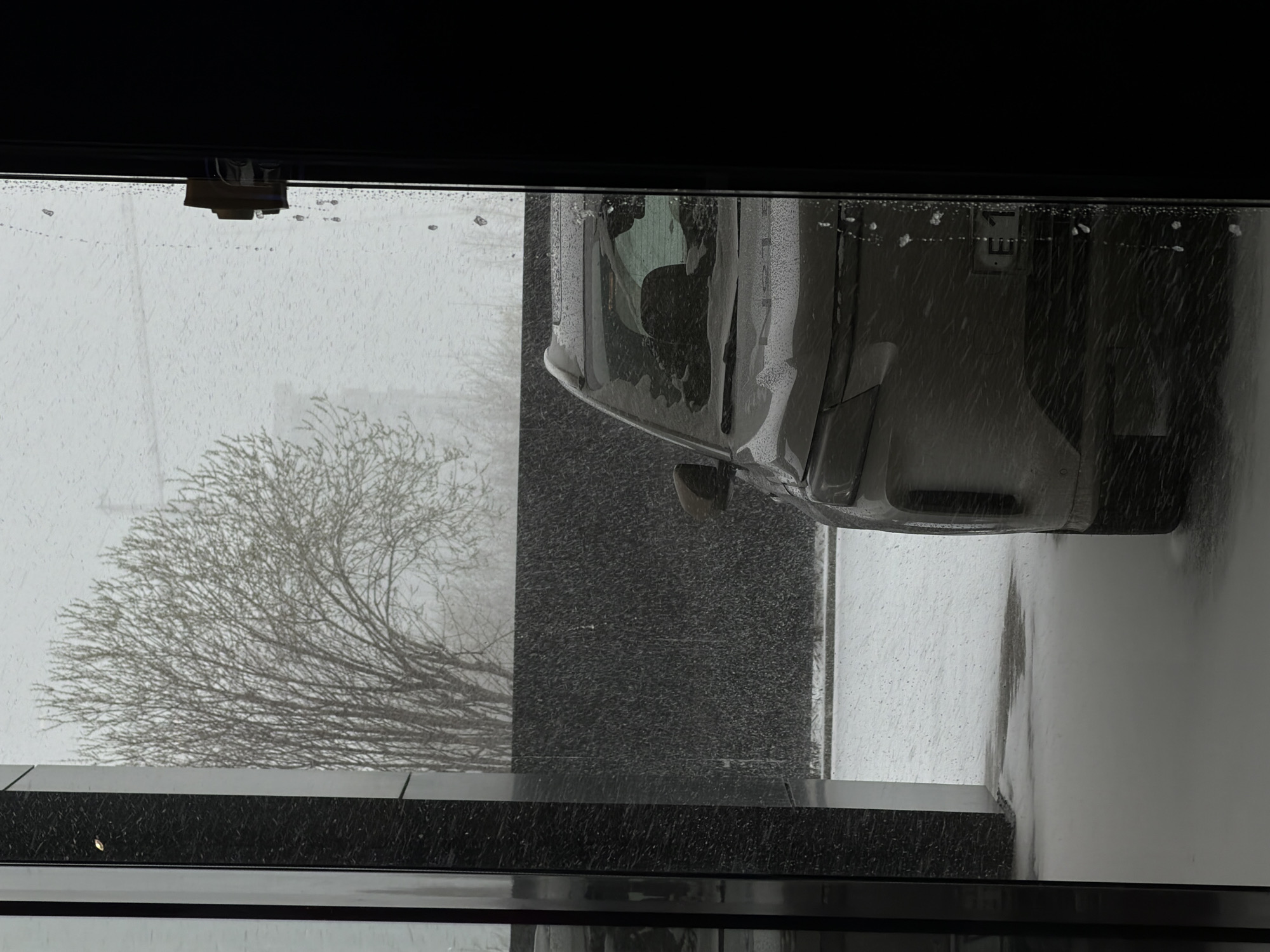 The image depicts a view from inside a building looking out onto a snowy scene. A car, partially covered in snow, is parked outside. A bare tree is visible to the left of the car. It is snowing heavily. The location is Izhevsk, Russia.