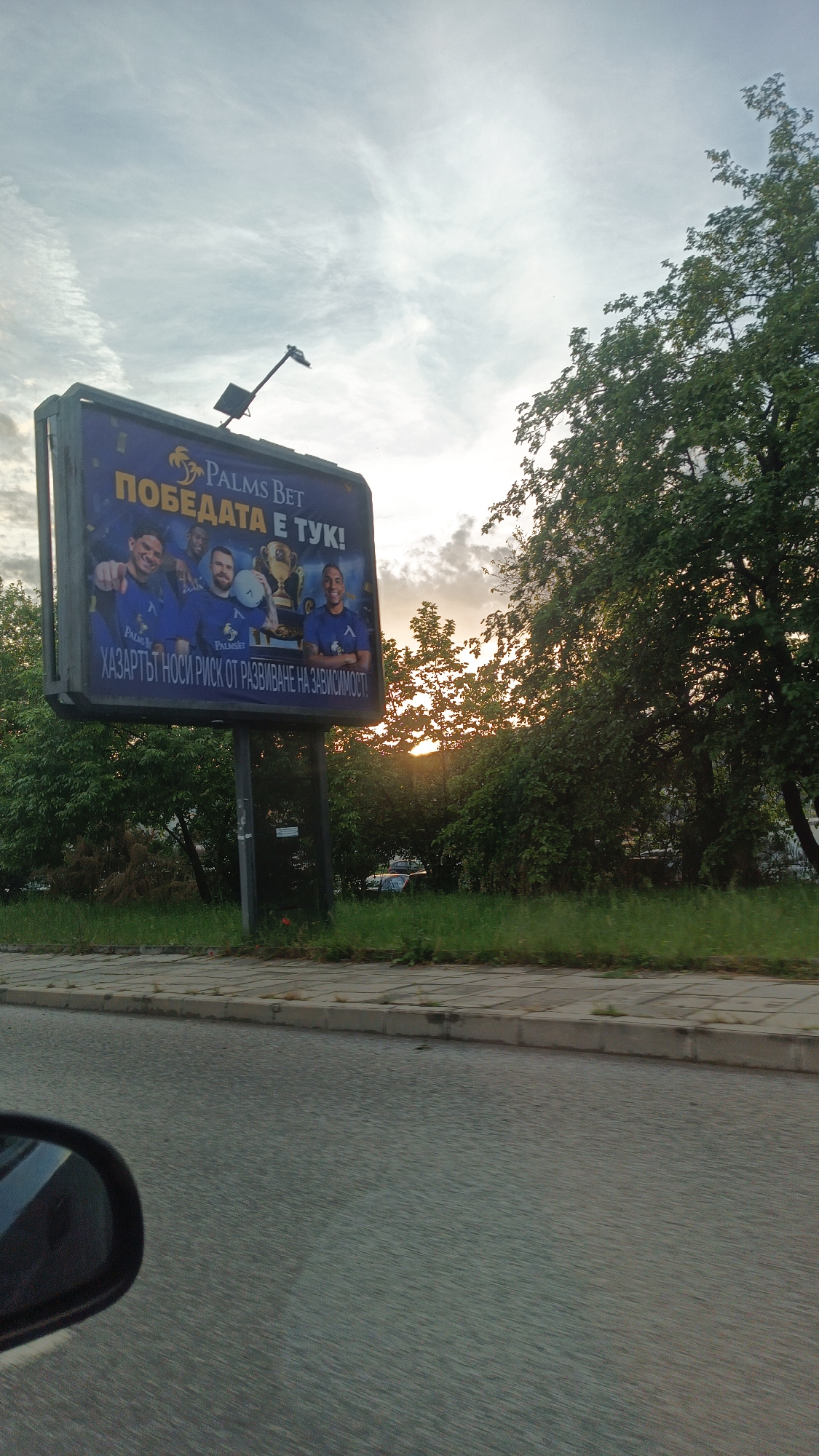 The image shows a billboard advertising "Palms Bet". The billboard features a picture of four men in blue shirts, one of whom is holding a trophy. The text on the billboard says "Palms Bet" and "Победата е тук!" (The victory is here!). It also includes a warning that "ХАЗАРТЪТ НОСИ РИСК ОТ РАЗВИВАНЕ НА ЗАВИСИМОСТ" (Gambling carries a risk of developing dependence). The billboard is located next to a street and there are trees and grass visible in the background. The lighting suggests it is either early morning or late evening. The location is the city of Burgas, Bulgaria. A portion of a car mirror can be seen in the lower-left corner of the picture suggesting the photo was taken from a moving car.