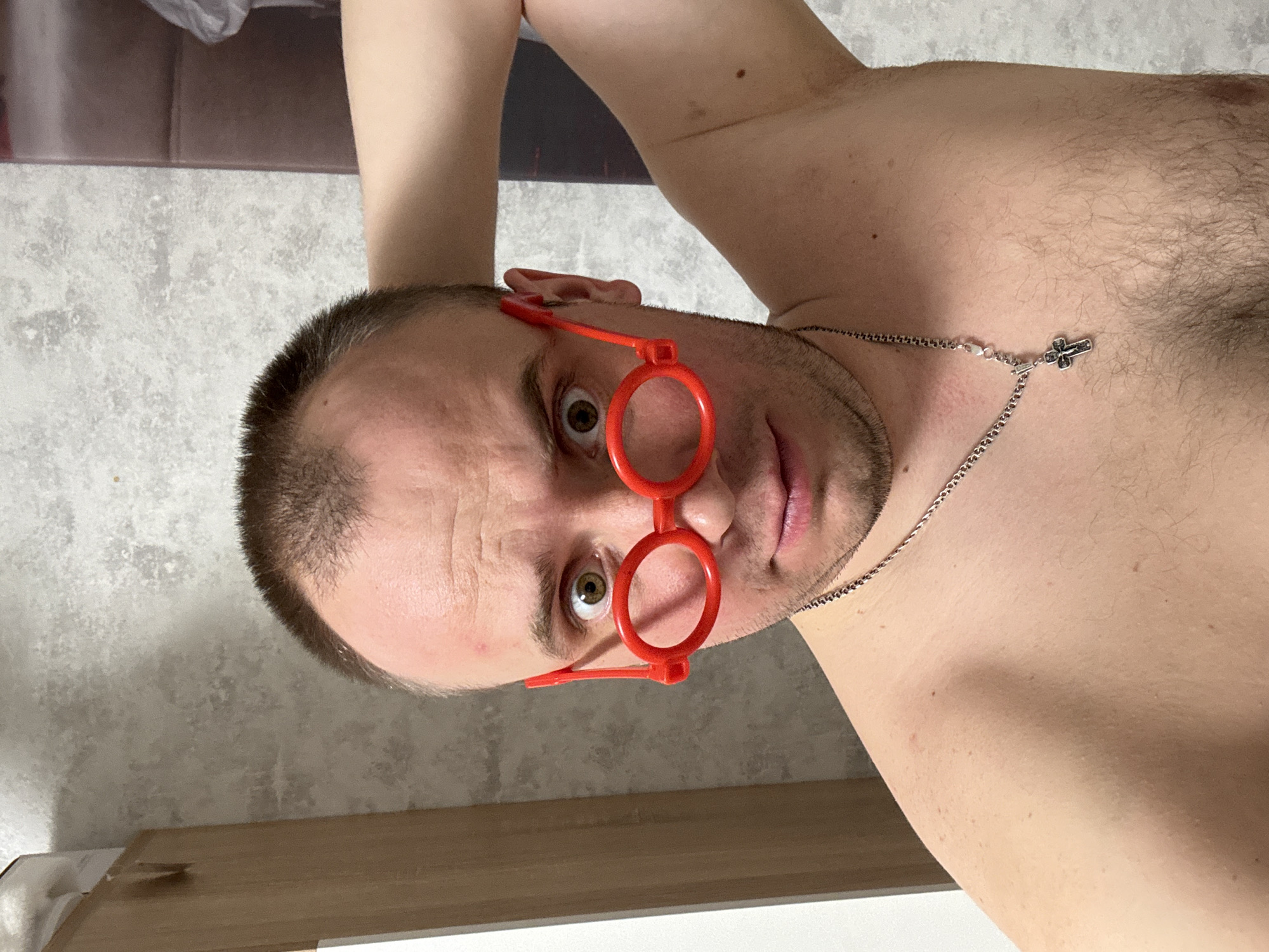 A shirtless man with short brown hair is wearing bright red, round novelty glasses and a silver necklace with a cross pendant. He is indoors. The background consists of a patterned wall, a brown surface, and a white surface. The location is unknown and cannot be determined from this image.