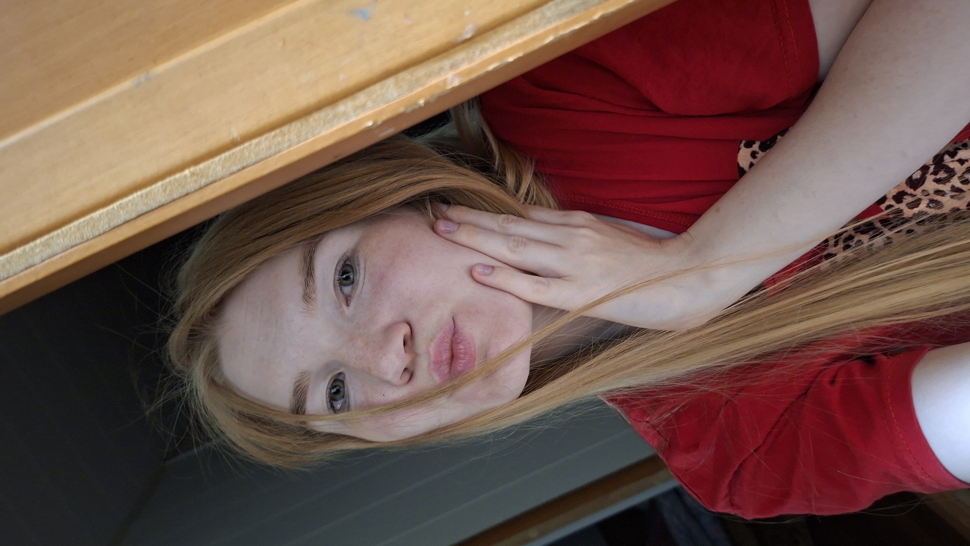 A young woman with long, blonde hair and fair skin with freckles is positioned in the foreground of the image, looking towards the camera. She has green eyes and her lips are pursed in a kissing gesture. Her right hand is placed on her left cheek. She is wearing a red t-shirt with a leopard print design visible on the right side of her chest. The background is dimly lit, with a dark wall on the left and a wooden structure on the right, suggesting an indoor setting. The overall lighting suggests natural light entering from the right side. The context of the city of Большой Букор, Russia is not evident from the visual information present in the image.