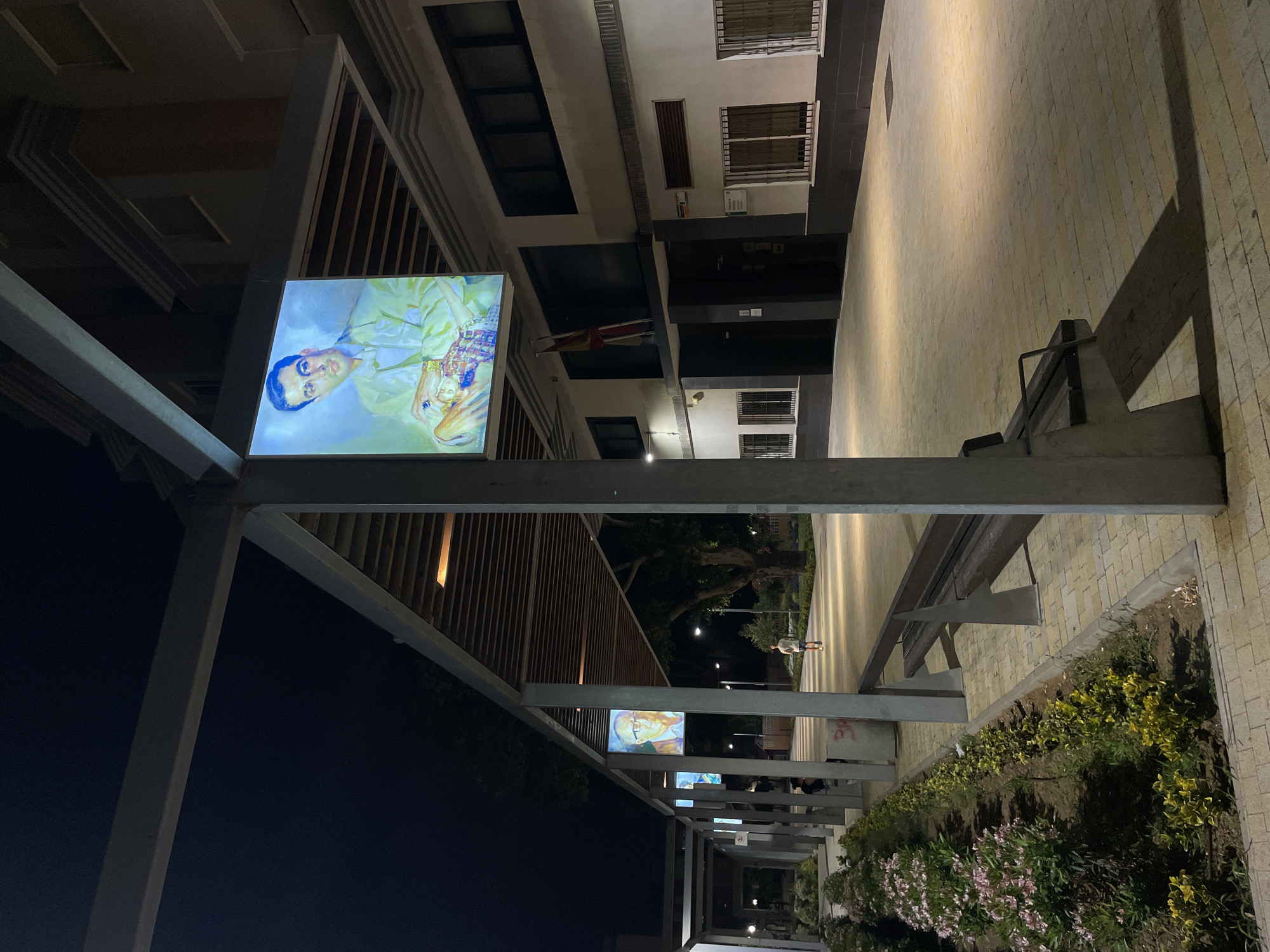 At night, a modern building with a white facade and dark trim stands illuminated by overhead lighting. The building has multiple windows with bars. A Spanish flag hangs on the facade near the entrance. In front of the building is a paved walkway bordered by planters with flowers and shrubs. A metal pergola runs along the walkway, supporting large illuminated portraits of men. One portrait, prominently displayed, shows a man in a light-colored suit holding a doll. Further down the walkway, another illuminated portrait of a man wearing glasses is visible. A solitary person is walking away from the camera down the walkway, and another person is seated under the pergola. Metal benches are positioned along the walkway. The overall scene suggests an outdoor public space at night, likely in La Línea De La Concepción, Spain, given the presence of the Spanish flag.