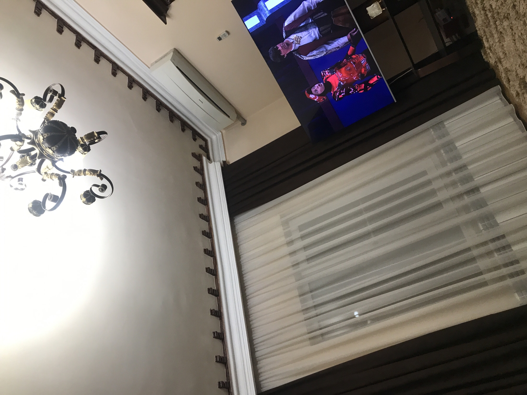 The image shows an interior view with a tilted perspective. A chandelier is visible in the lower-left corner against a white wall with decorative molding. A white air conditioning unit is mounted on the wall near the top. A television screen displaying two people in costume is visible in the upper-right. The scene includes a window covered by light-colored horizontal blinds. Brown curtains frame the window. The location is not possible to determine from this image.