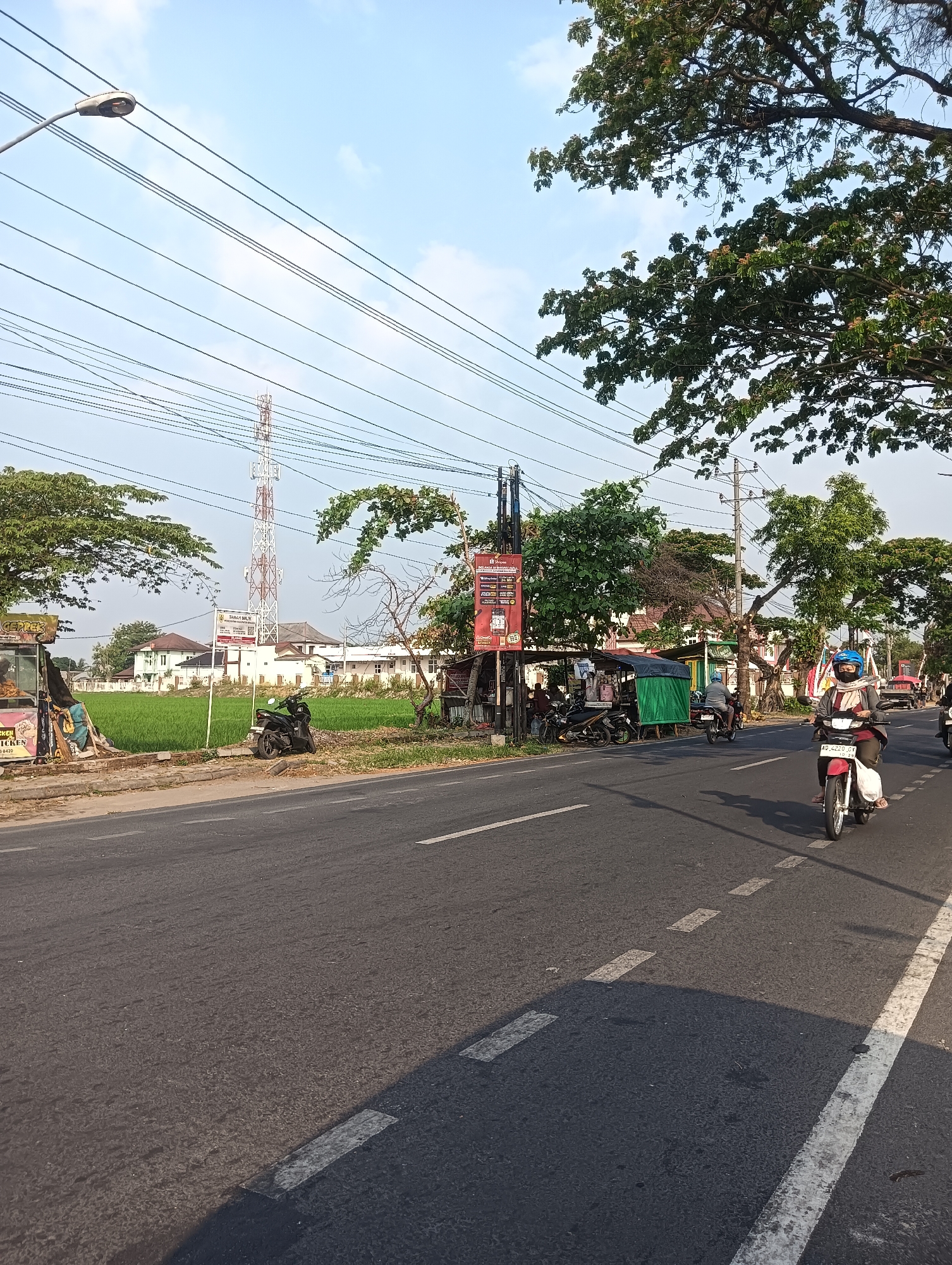 This is a street-level, eye-level, medium shot of a roadside scene in Sragen, Indonesia. The image captures a paved road with clear white lane markings, a slight shimmer on the asphalt suggesting a warm day. On the right side of the road, a woman wearing a helmet and a light-colored headscarf rides a red and white motorcycle, her face partially obscured by a dark mask. Her shadow is cast across the road. Further down the road, several other motorcycles are parked.
To the left of the road, a green field stretches behind a low barrier. A tall, red and white lattice communication tower is prominent in the background. Several low buildings, likely residential or commercial, are situated behind the field. A street lamp is visible on the left, silhouetted against a clear blue sky with some wispy clouds. Trees with dense green foliage line parts of the roadside, and a banner with text is displayed vertically on a pole in front of some stalls. The atmosphere appears to be during the daytime, with sunlight casting distinct shadows.