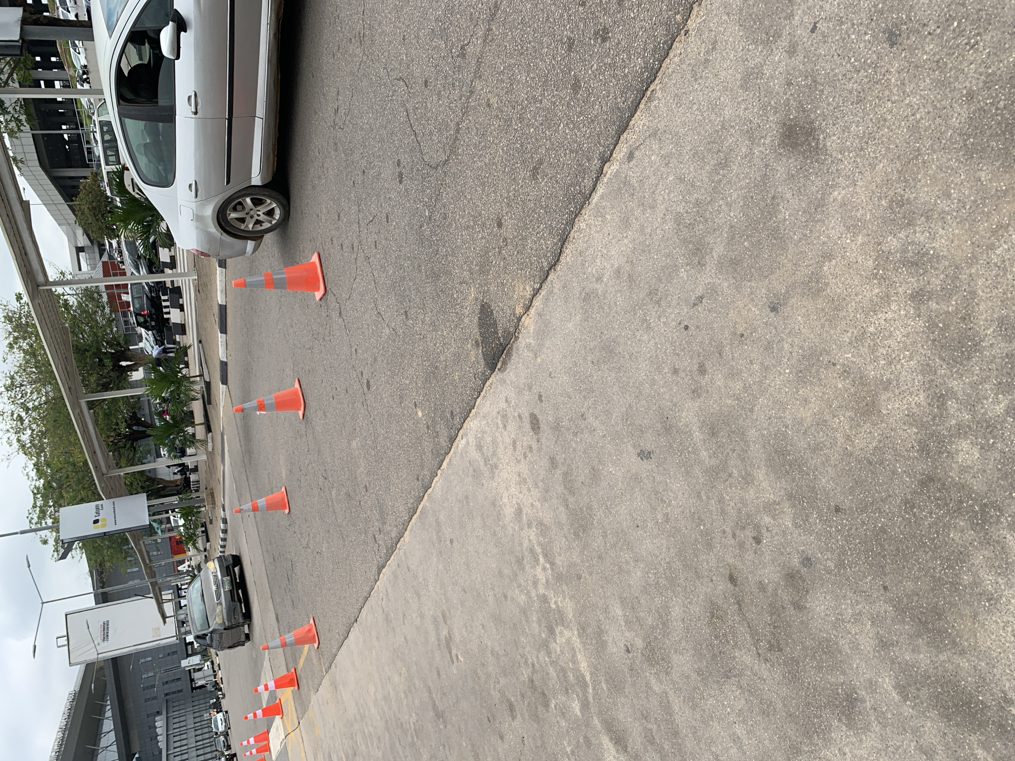 The image depicts a bright, sunny daytime scene on a roadside in Gwaska, Nigeria. A light-colored four-door sedan, possibly white or silver, is parked or slowly moving on the left side of the frame, positioned on an asphalt road. A distinct line of orange and white traffic cones stretches diagonally from the foreground to the mid-ground, serving as a barrier or demarcation along the edge of the asphalt. These cones separate the main roadway from a lighter, grey concrete surface, likely a sidewalk or shoulder, which occupies the right half of the image.
Further down the road, beyond the first few cones, a dark-colored sedan with noticeable damage to its front bumper is visible. In the background, tall modern buildings with glass windows and some scaffolding can be seen under a clear sky. Lush green trees and various plants line the road and buildings. Multiple streetlights and poles are scattered throughout the scene.
Several billboards are visible in the background. One prominently displays the text "Tatum Homes" along with a partial website address starting with "www.tatumho...". Another billboard features phone numbers beginning with "+234" and what appears to be text related to property sales. A sign displaying "Lagos" is also identifiable. The strong sunlight casts clear shadows, indicating a bright and clear day. The overall setting suggests a busy urban or commercial area, possibly near an airport given the architectural style and infrastructure.