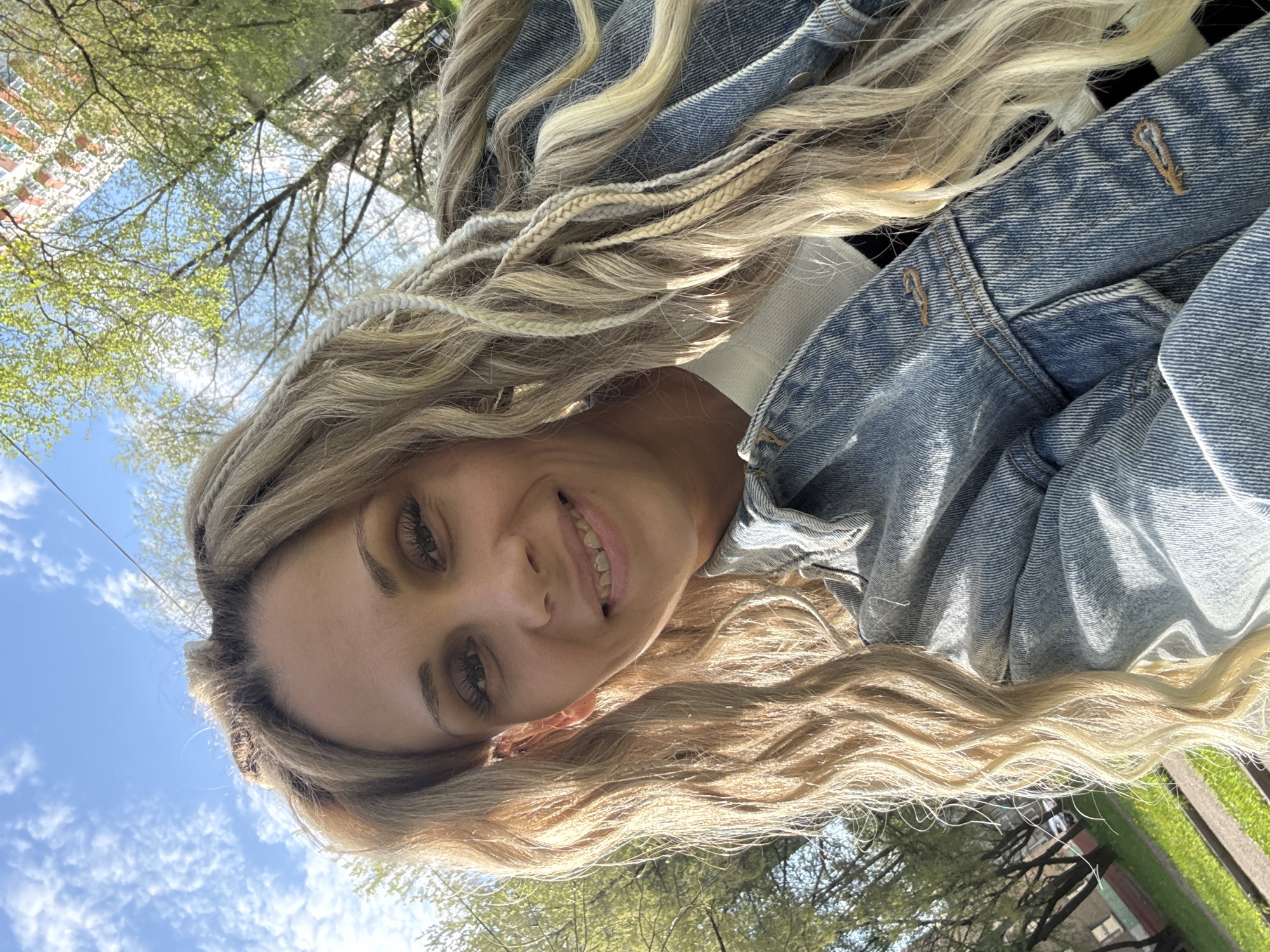 The image is an upward-angled selfie featuring a woman with light blonde, wavy hair and several small braids. She is smiling. She is wearing a blue denim jacket. Behind her are trees with light green leaves, a blue sky with some clouds, and the top of a red and white building. The location is Rusanovo, Russia.