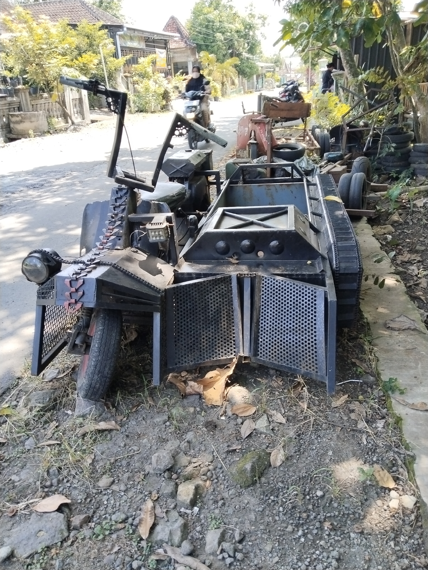 The image shows a custom-built vehicle with a tank-like appearance parked on the side of a road in Dawungan, Indonesia. The vehicle has a black frame with a grill-like front, three tires, and a chain track on the right side. It has unique handlebars and a headlight. It is parked next to a concrete curb with some dirt and debris around it. A person on a motorcycle is traveling down the street away from the camera. Another person is nearby. Additional vehicles and objects are partially visible in the background. Houses and trees are also visible along the street.