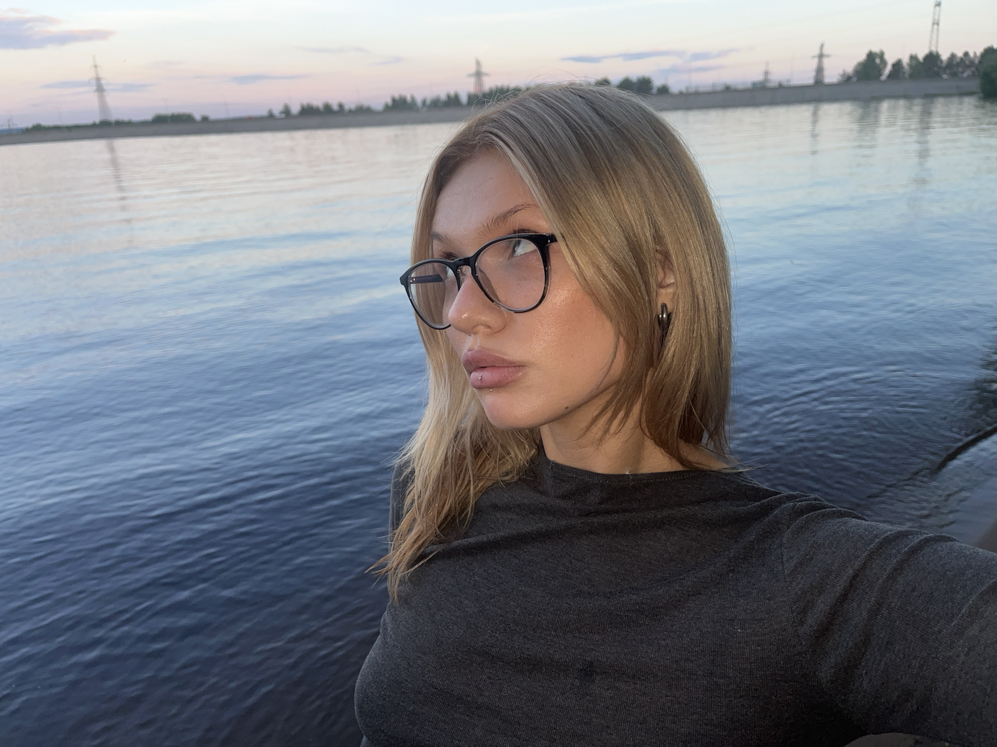 A young woman with blonde hair, fair skin, and black-rimmed glasses is the central subject of this outdoor photograph. She wears a dark grey or black long-sleeved top, a small silver labret piercing below her lower lip, and a dark hoop earring in her left ear. She is captured in a half-profile pose, looking thoughtfully towards the upper left of the frame, with a serene and pensive expression.
The setting places her beside a large body of water, likely a river or lake, which occupies the majority of the background. The water's surface shows gentle ripples and reflects the soft, diffused light of the sky. In the distant background, across the water, a low-lying shore or embankment is visible, dotted with sparse trees or vegetation and several industrial structures, including power lines and a prominent tall power tower.
The lighting suggests late afternoon or early evening, with the sky displaying soft pastel hues of pale pink, purple, and blue, indicative of either sunset or twilight. The overall atmosphere is calm and peaceful. No visible text is present in the image. The scene offers a view of a natural waterfront area bordering a landscape with industrial elements, set in a location such as Noviy, Russia.