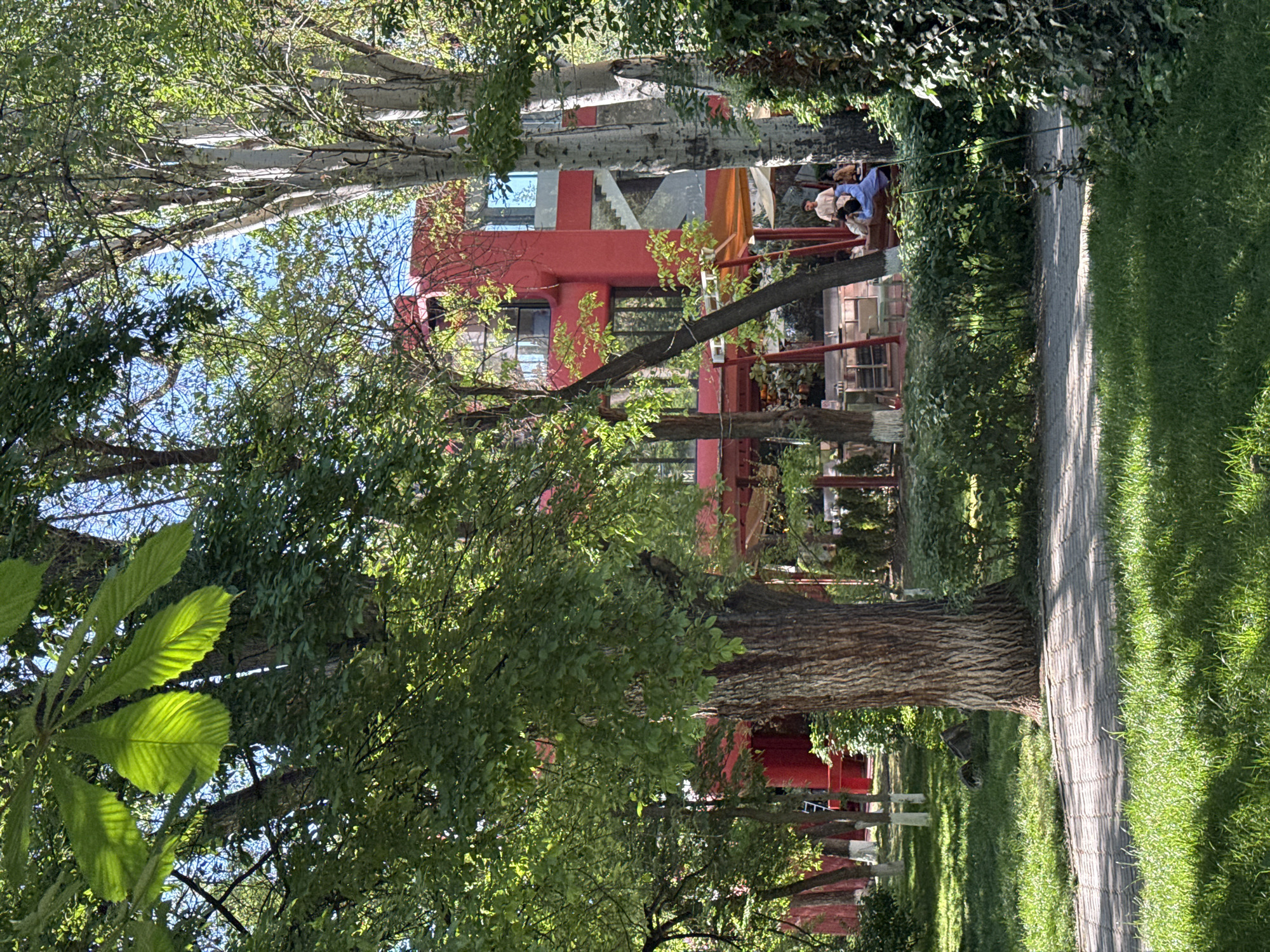 The image shows a park scene in Yerevan, Armenia, featuring a striking red building amidst lush greenery. A paved pathway runs along the right side of the frame, bordered by grass and trees. The red building is partially obscured by trees, but its modern architecture, including glass windows and exterior staircases, is visible. People are present on the building's exterior, seemingly engaged in conversation. Trees with thick trunks and abundant foliage dominate the foreground, casting shadows across the scene. The overall impression is that of a vibrant urban oasis.