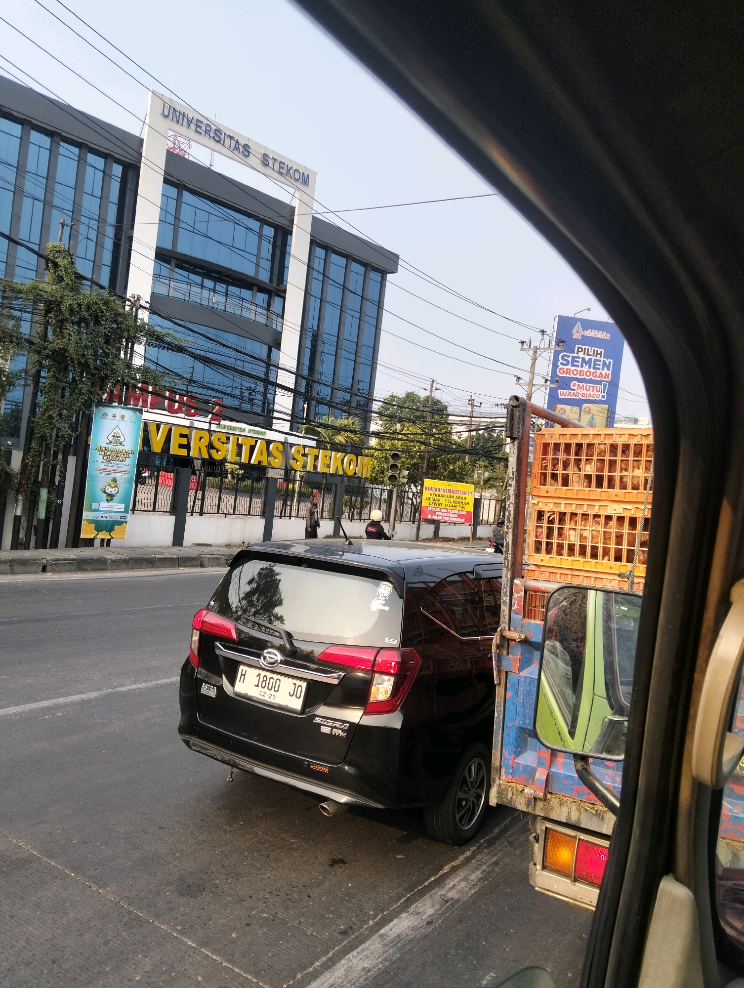 This is a street-level, medium-angle, outdoor, daytime shot taken from inside a vehicle looking forward and slightly to the right. The primary focus is on a black Daihatsu Sigra car with the license plate "H 1800 JO". Behind and to the right of the Sigra is a blue truck with a distressed paint job carrying stacked orange crates filled with live chickens.
In the background, a modern, multi-story building with a dark facade and blue glass windows is visible. A large sign above the entrance reads "UNIVERSITAS STEKOM". To the left of the building, a tall, vertical banner is displayed. Further down the street, a traffic light is visible, and a few people, some on motorcycles and one standing, are present. A billboard with Indonesian text, including the words "PILIH SEMEN GROBOGAN", is also visible in the distance.
The scene appears to be a busy urban street in Semarang, Indonesia, likely during daylight hours given the illumination. The overall impression is of a traffic scene with urban elements and commercial advertising.