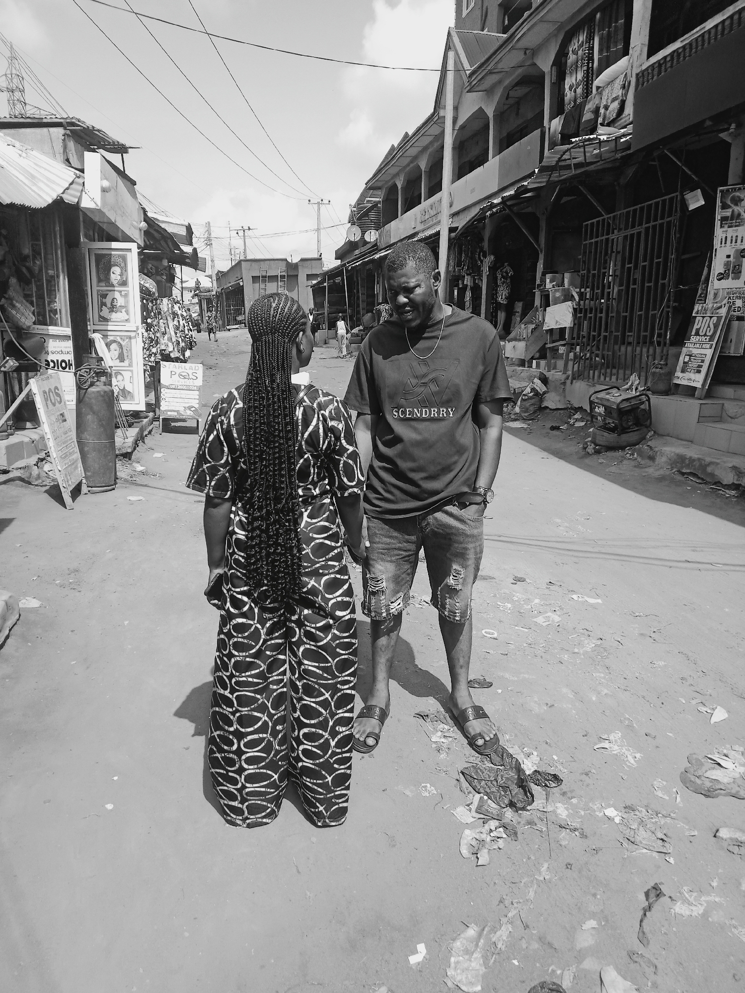 Two individuals, a man and a woman, stand on a dirt road in what appears to be a bustling commercial area. The woman faces away from the viewer, showcasing her long braided hair and patterned clothing. The man, wearing a t-shirt with "SCENDRRY" printed on it and ripped denim shorts, looks down at her.
The setting is a street lined with shops, some with corrugated metal roofs, others with more permanent structures. Signboards advertising services like "POS" (Point of Sale) are visible. Debris litters the ground. Overhead, power lines crisscross the sky. This appears to be the city of Onitsha, Nigeria.