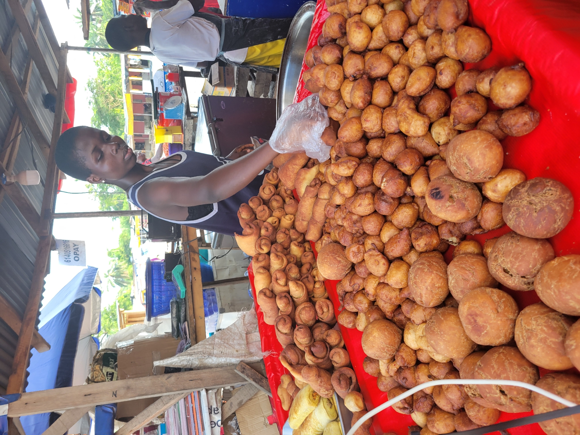 A young woman, wearing a dark blue tank top, stands behind a red tablecloth laden with fried pastries in a market stall in Egbagu, Nigeria. Her right hand, encased in a clear plastic glove, reaches towards the pastries. The pastries are varied in shape and size, with some resembling small, round balls and others appearing as rolled or coiled dough. In the background, a young boy in a white t-shirt and an apron stands near some containers. The stall is covered by a corrugated metal roof supported by wooden beams, and a blue and white umbrella is visible overhead. Various items, including boxes, blue plastic baskets, and what appears to be a weighing scale, are on shelves behind the woman. The scene suggests a bustling outdoor market environment.