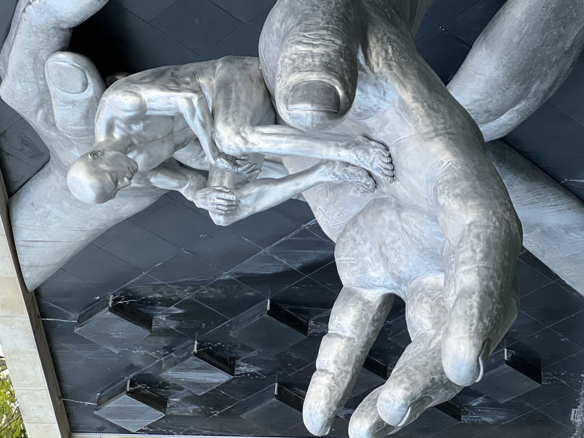 The image depicts a large, metallic sculpture of a man sitting in a pensive pose on two cupped hands. The sculpture is situated in front of a black, diamond-patterned wall with water cascading down it from multiple outlets. The man is bald, muscular, and appears to be contemplating something, with his head bowed and his hands resting on his knees. The hands supporting him are also metallic and detailed, with visible fingernails and skin texture. The overall impression is one of contemplation and support, with the water feature adding a dynamic element to the scene. The location is identified as Krasnodar, Russia, however, this information is not visually verifiable from the image.