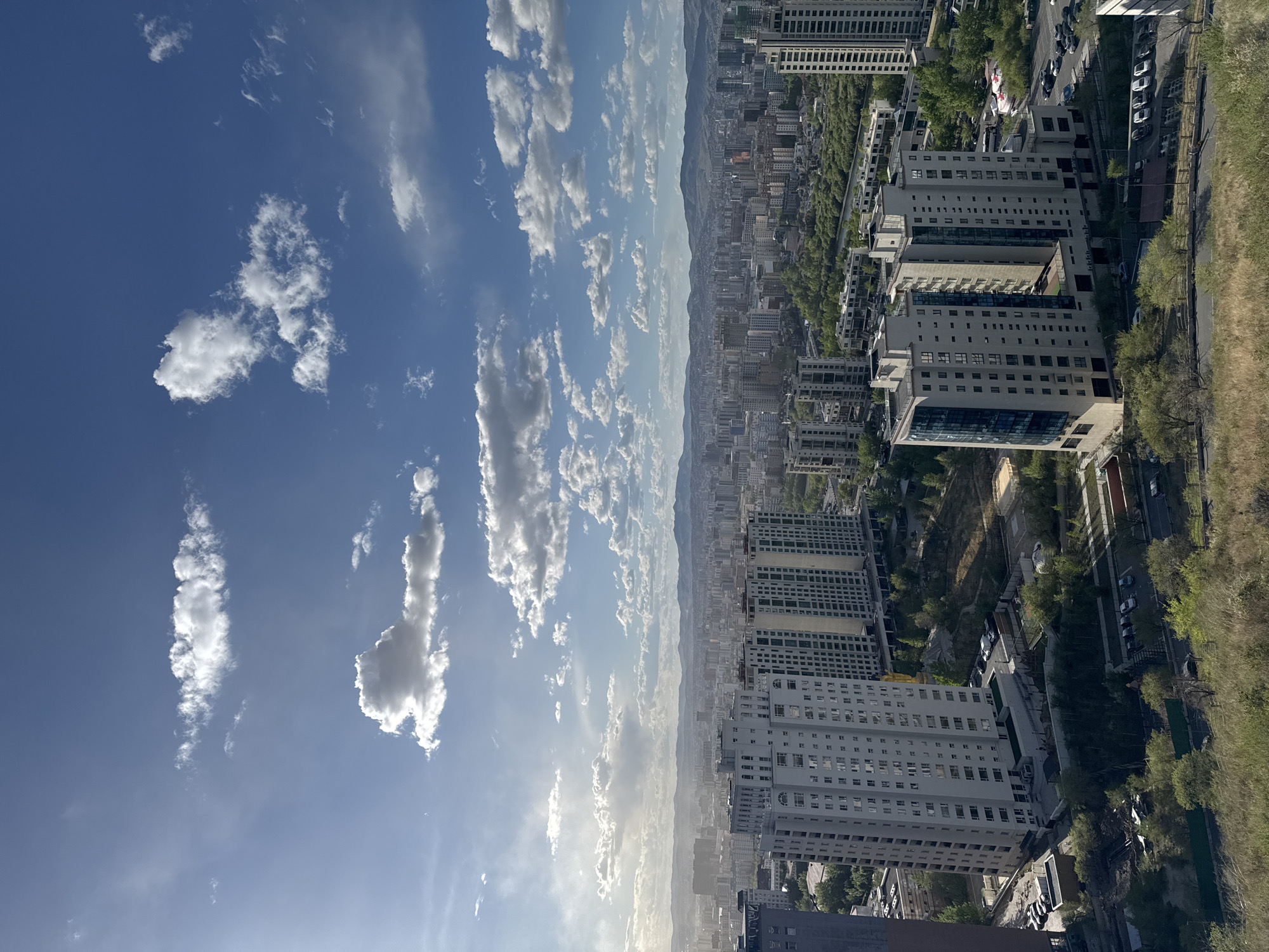The image is an elevated view of Ulaanbaatar, Mongolia. In the foreground, there are several multi-story buildings, likely apartment complexes. Adjacent to these buildings, a road with parked cars is visible, bordering a green, tree-filled area. Beyond the buildings and greenery, the cityscape of Ulaanbaatar extends toward a distant mountain range. The sky above is blue with scattered white clouds.