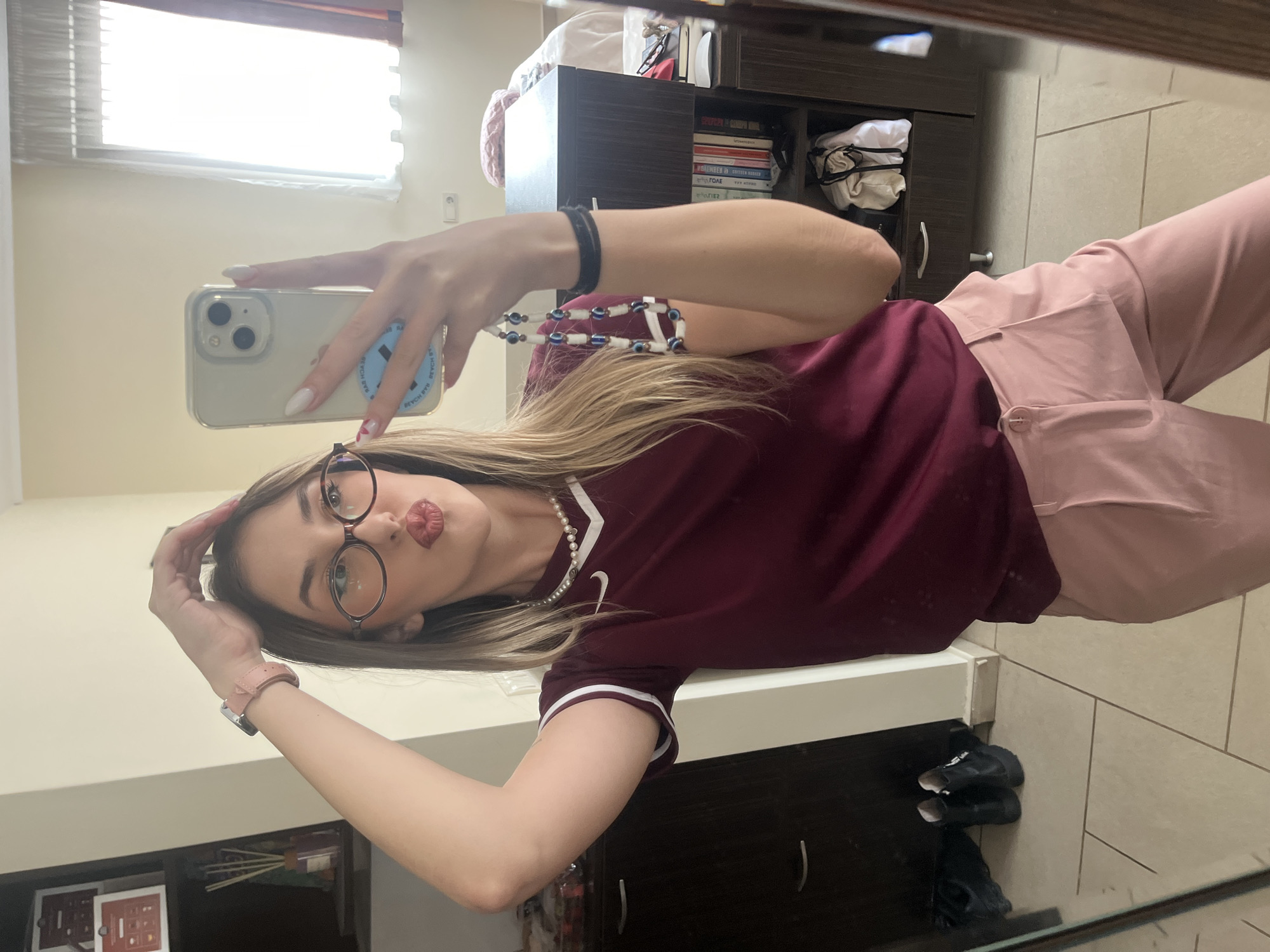 A young woman is taking a selfie in a mirror. She is wearing glasses, a maroon shirt with white trim, pink pants, a pearl necklace, and a bracelet. She is holding a white phone with a clear case. Behind her is a dark brown cabinet with books and white items. The floor is tiled. A window is visible with blinds. It is not possible to determine if this image was taken in Sofia, Bulgaria.