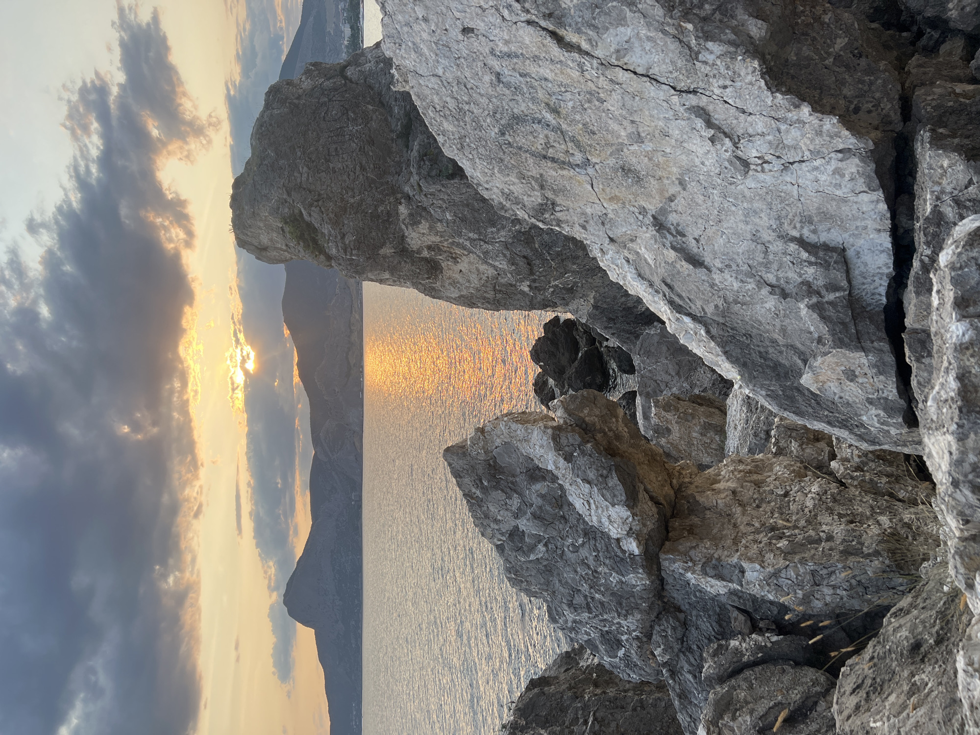 The image depicts a rocky coastline at sunset near Sudak, Ukraine. The foreground is dominated by large, grey, jagged rocks, with sparse dry grass growing between them. Beyond the rocks, the calm sea reflects the warm orange and yellow hues of the setting sun. In the background, a range of low, silhouetted mountains forms the horizon, with a hint of a town or settlement visible on the right side. The sky above is dramatic, with dark, textured clouds partially obscuring the sun, allowing beams of light to break through and illuminate the scene. There are no people or animals visible in the image.