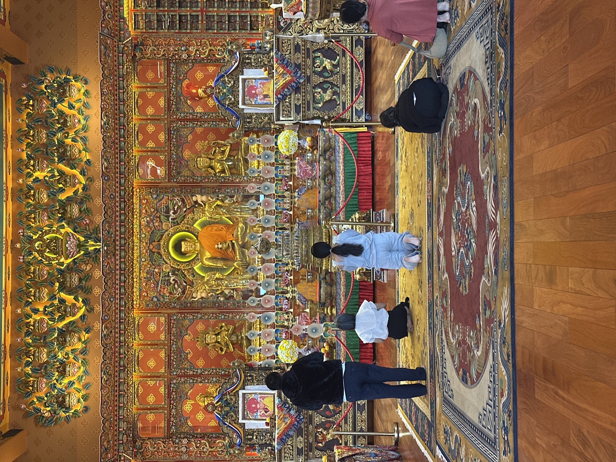 The image depicts the interior of a Buddhist temple in Bookabang, Vietnam, with several individuals engaged in prayer. In the foreground, a large, ornate rug with a dragon motif covers the wooden floor. Several people are seen kneeling and praying on the rug, facing a richly decorated altar.
The altar features a central golden Buddha statue, illuminated with a halo. Flanking the Buddha are smaller golden statues and intricate wall hangings. Several framed portraits, likely of spiritual leaders, are also displayed. Decorative elements, including colorful fabrics, offerings, and floral arrangements, adorn the altar and surrounding structures. The temple walls and ceiling are elaborately decorated with gold and vibrant colors, featuring intricate carvings and patterns. The overall atmosphere suggests a place of worship and spiritual devotion.