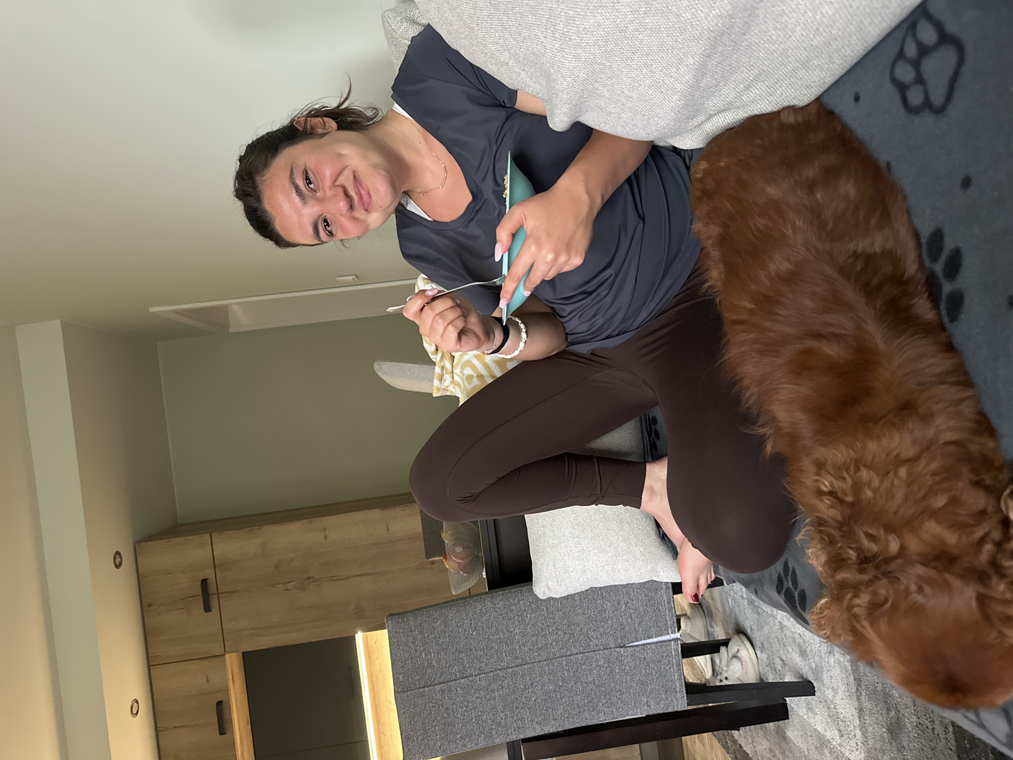 The image appears to be taken indoors, possibly in a living room or similar domestic space. A woman with dark hair pulled back is seated on a sofa, holding a blue bowl and a fork, seemingly in the act of eating. She wears a dark grey top and brown leggings, and several bracelets are visible on her wrist. Her posture suggests she is leaning against the back of the sofa. To her right is a brown dog, possibly a poodle, sitting beside her on the sofa. The room is furnished with a grey chair and a wooden cabinet on the left side of the frame. The environment appears relaxed and informal. The location is not identifiable as Sofia, Bulgaria, from visual cues within the image.