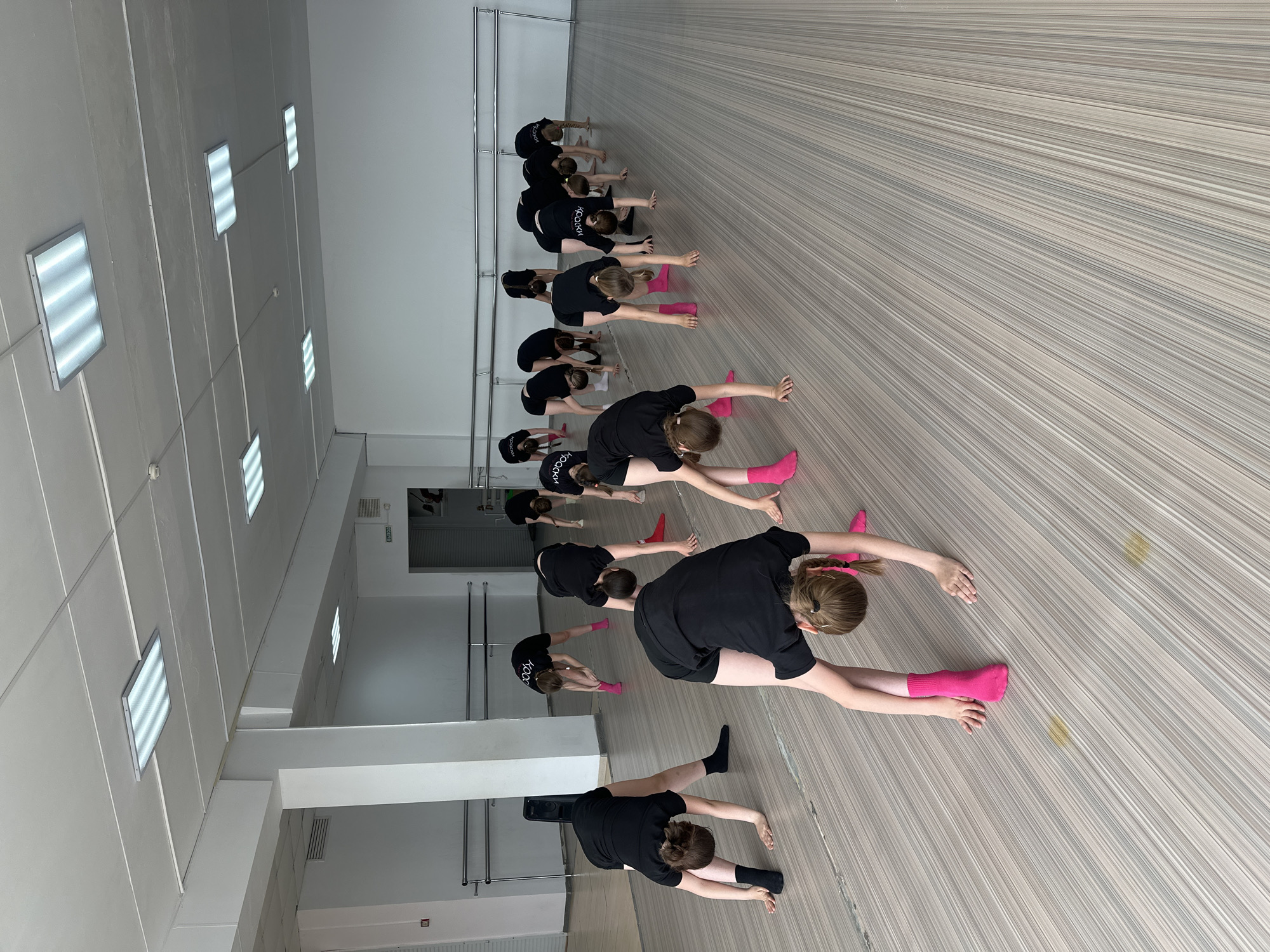 A group of approximately a dozen young women are stretching in a dance studio. They are lined up in rows, all wearing black tops and shorts, with some wearing bright pink socks and others black. The room is brightly lit with panel lights and has a wooden floor. There is a metal bar along one wall, and a doorway in the back of the room. The location is a dance studio in Votkinsk, Russia.
