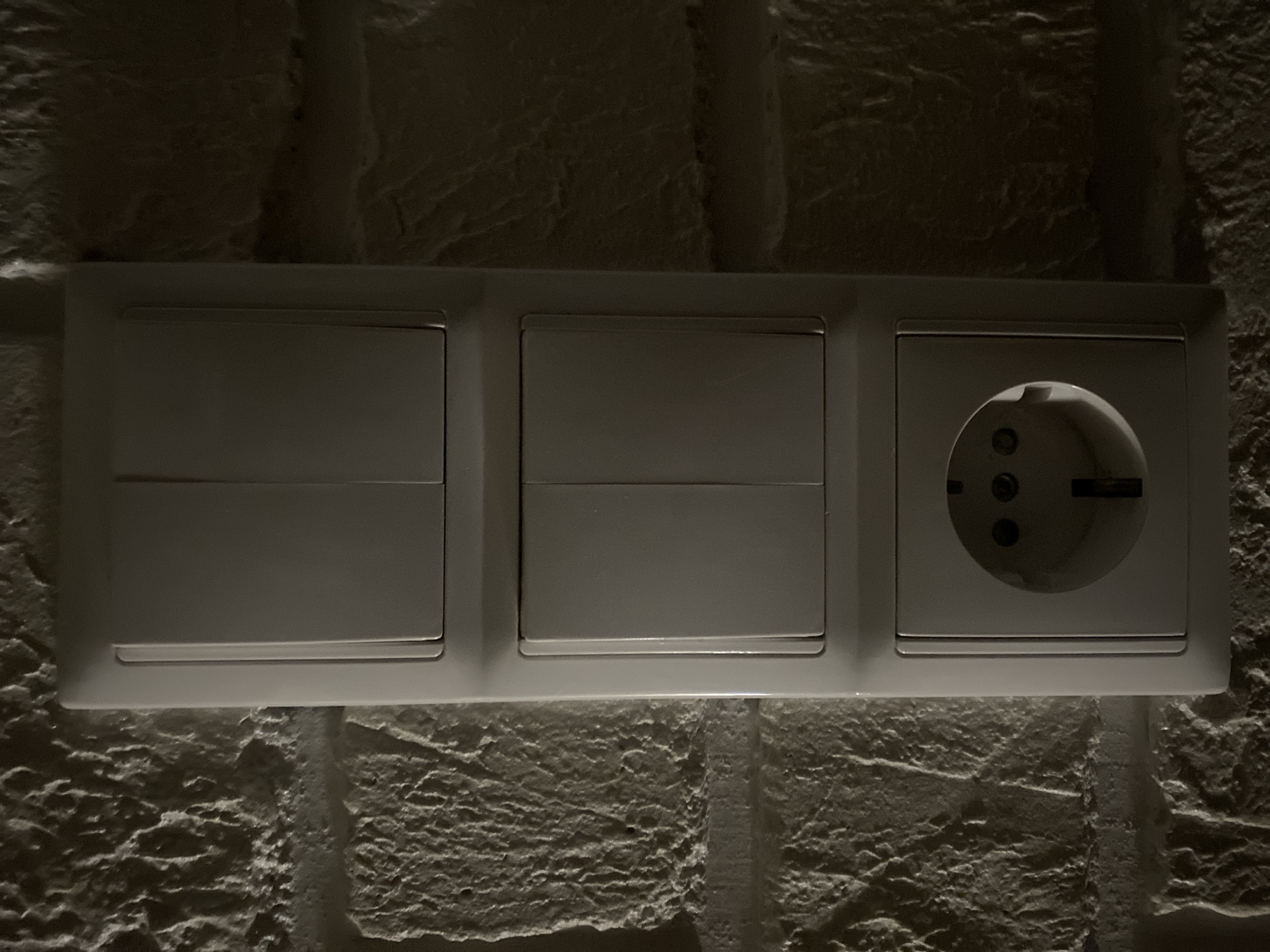 A close-up, eye-level, medium shot shows a vertical electrical outlet and two rows of light switches mounted on a textured, light-colored stone wall. The outlet is at the bottom of the frame, a standard European-style receptacle with three circular openings and a grounding pin slot. Above the outlet are two stacked electrical switch modules. The top module contains two light switches, and the module below it also contains two light switches. The switches and outlet are all housed within a single white plastic plate. The wall behind the electrical fixture is made of rough-hewn stones, creating a contrast in textures between the smooth plastic and the rough stone. The lighting is dim, with shadows highlighting the texture of the stone wall and creating a subdued atmosphere. The location is not specified beyond "Unknown, Unknown."