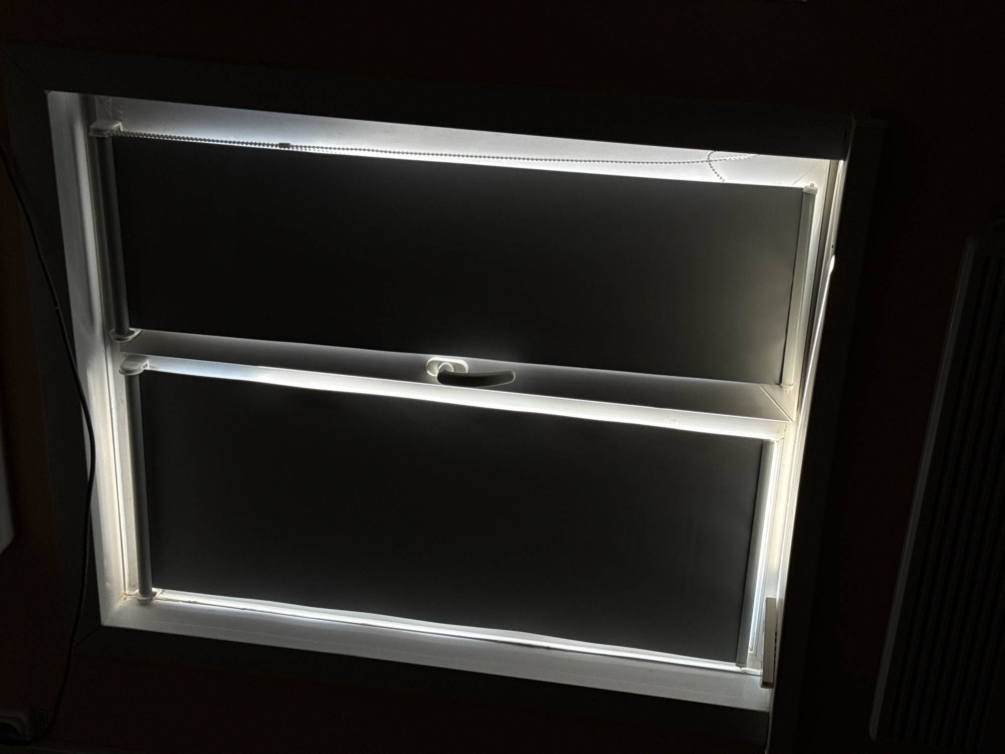 The image shows a closed window with roller blinds. Light streams in from the left, illuminating the edges of the blinds and the window frame. The window is divided into two panes by a vertical mullion. A handle is visible on the right pane. The roller blinds are dark grey. Below the window is a radiator. The scene is indoors, with dark walls. The location is specified as Қопа ауылдық округі, Kazakhstan, implying a rural or semi-rural setting. No people are visible. The overall impression is one of quiet domesticity or an empty room.