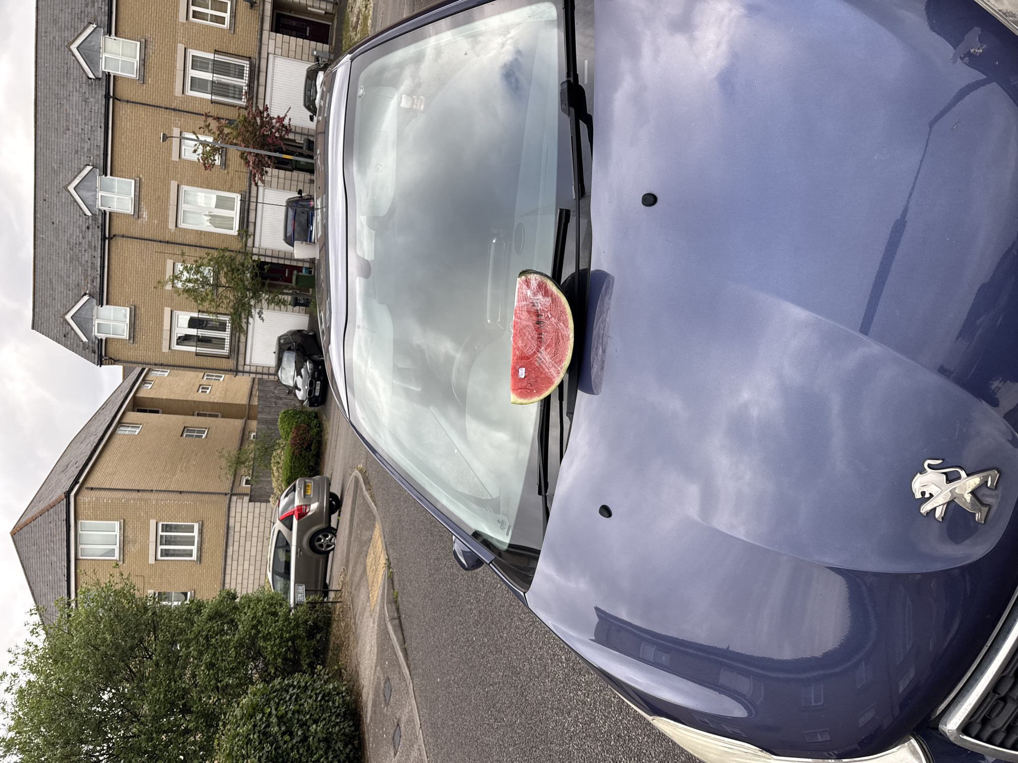 A slice of watermelon rests on the windshield of a blue Peugeot automobile parked on a residential street in what appears to be Great Yarmouth, United Kingdom. The car is oriented diagonally in the frame, with its front facing the viewer. Behind the car, typical British terraced houses are visible, constructed from yellow brick with gray tiled roofs. A silver SUV is parked further down the street. The sky is overcast, suggesting a cloudy day. No people are visible in the shot.