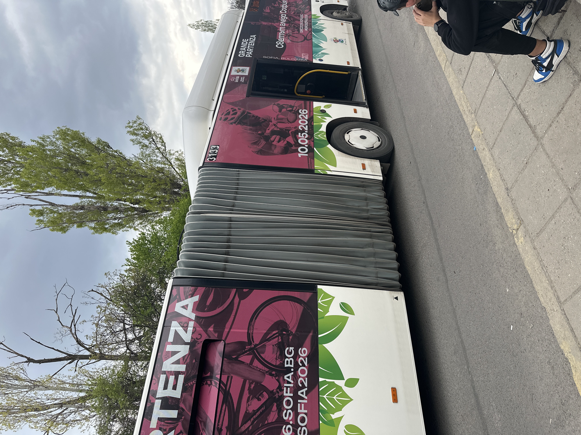 An articulated bus is prominently displayed on an asphalt road in Sofia, Bulgaria, under an overcast sky. The bus features a distinct white, maroon/pink, and green promotional wrap advertising "GRANDE PARTENZA" and "GIRO SOFIA 2026," a cycling event slated for "10.05.2026." The wrap showcases graphics of a cyclist in action and bicycles, complemented by green leaf designs, symbolizing perhaps an environmental aspect or the start of the event.
Visible text on the bus includes "SOFIA, BULGARIA," the Bulgarian phrase "Светът гледа София" (The world watches Sofia), "GIRO2026.SOFIA.BG," and the hashtag "#GIROSOFIA2026." The bus also displays the identification number "3133" near its flexible, accordion-like mid-section.
On the adjacent concrete sidewalk, a person is seen squatting or sitting. They are dressed in a black hoodie, black pants, and distinctive blue and white high-top sneakers. A dark helmet or cap rests near their head. In the background, trees with lush green foliage are visible, indicating an outdoor, urban setting. The cloudy sky suggests a mild, possibly spring or autumn, daytime atmosphere. The scene captures a moment of urban life where public transport doubles as a promotional platform for a future major sporting event.