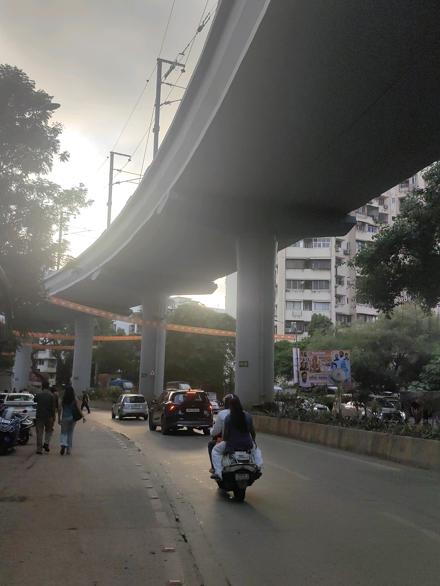 The image depicts a street scene in Mumbai, India, likely during the late afternoon or early evening, given the low angle of the sun and long shadows. The prominent feature is a large, elevated structure, possibly a flyover or metro line, supported by concrete pillars. The sky is hazy with bright sunlight filtering through.
In the foreground, a road with traffic is visible. Two people are walking away from the camera on the left side of the road. Further ahead, a black SUV with its brake lights illuminated is in motion. On the right side of the road, a scooter with two occupants, a driver and a pillion rider, is also moving forward. The pillion rider is wearing a dark blue top and the driver is wearing a white garment. Several other vehicles, including cars and a scooter, are present on the road.
Along the side of the road, there is greenery and a tall building complex with multiple floors and windows. A large billboard or banner with images of people and text in an Indian script is displayed. Orange and green decorative streamers are hanging from the pillars supporting the elevated structure. The overall environment suggests an urban setting with ongoing traffic and pedestrian activity.