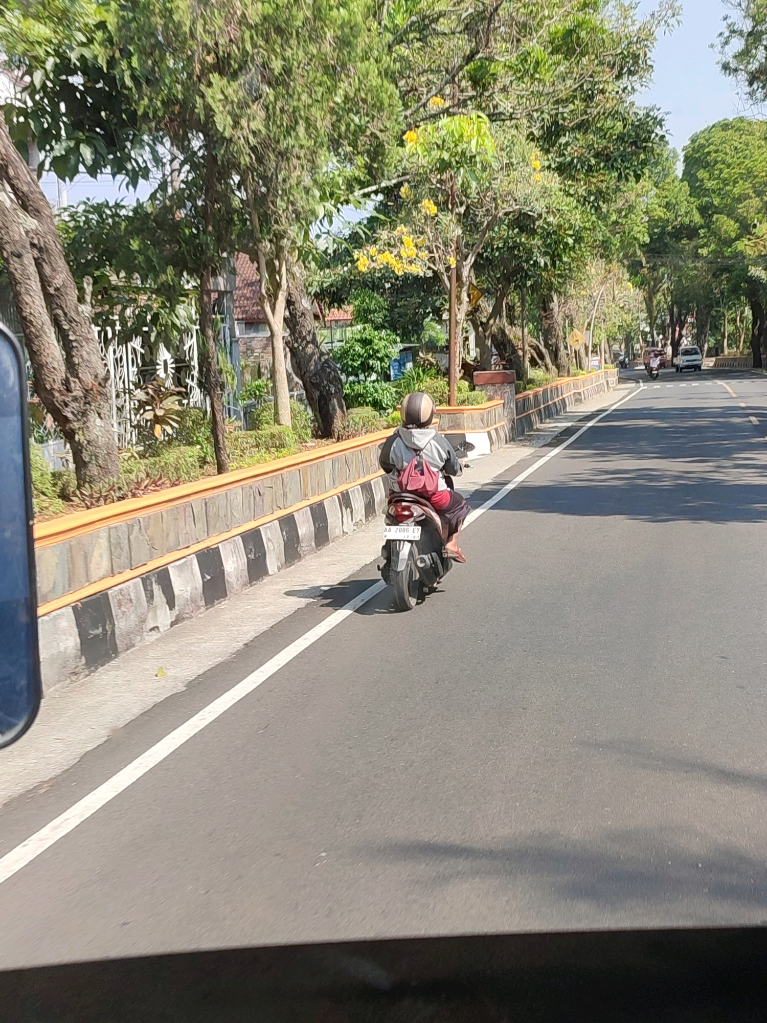 The image depicts a street scene in Kedu, Indonesia, with a person riding a scooter on the right side of the road. The scooter rider is wearing a helmet and a red backpack. The road is paved with asphalt and has a white line marking the lane. To the left of the road, there is a sidewalk and a low wall made of stone and concrete. Beyond the wall, there are trees with green foliage and some yellow flowers. The sky is clear and blue. In the background, there are buildings and other vehicles on the road. The overall scene suggests a typical day in a city or town.